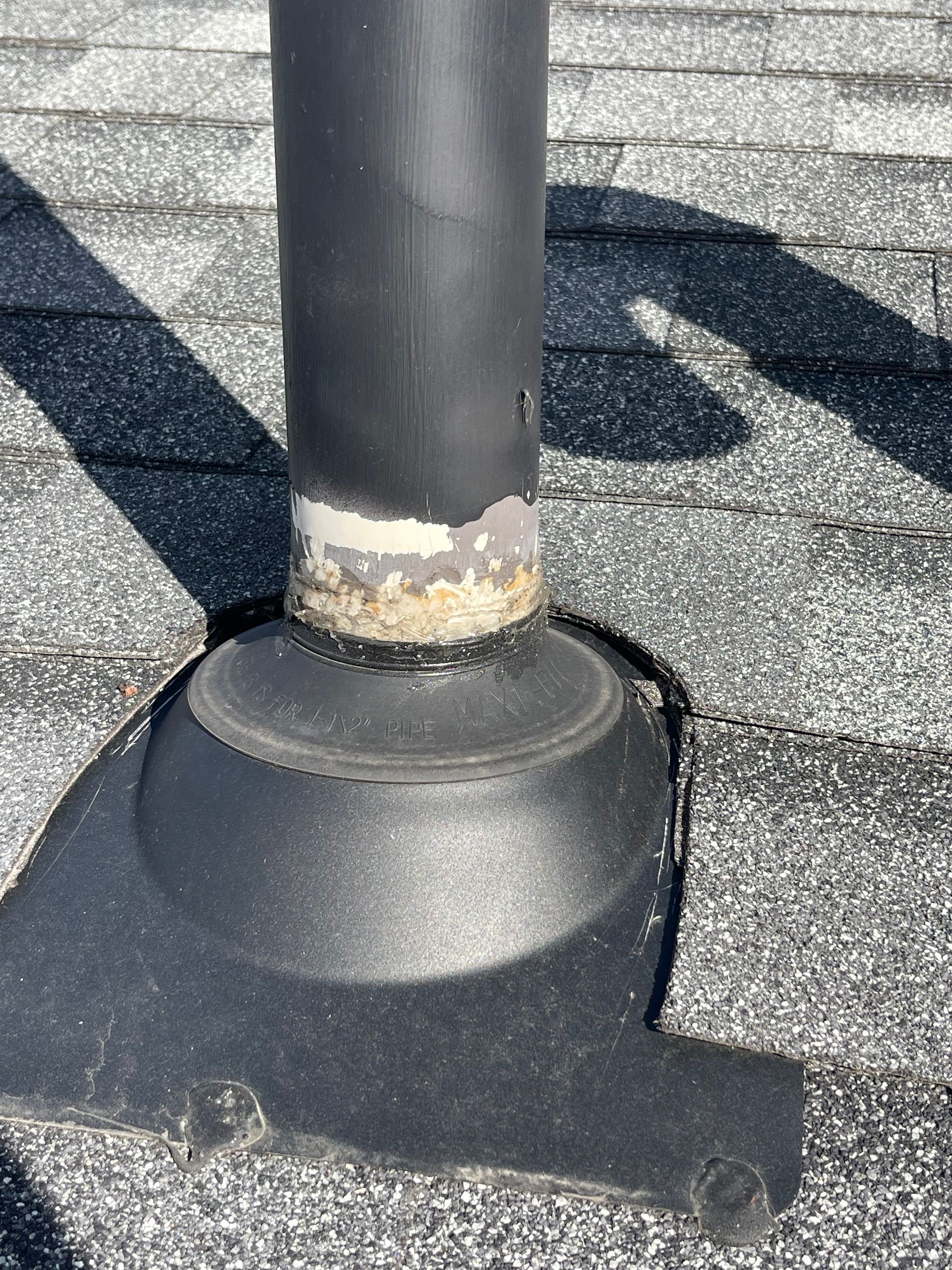 Roof vent pipe with black flashing on gray asphalt shingles, showing weathered seal at the base