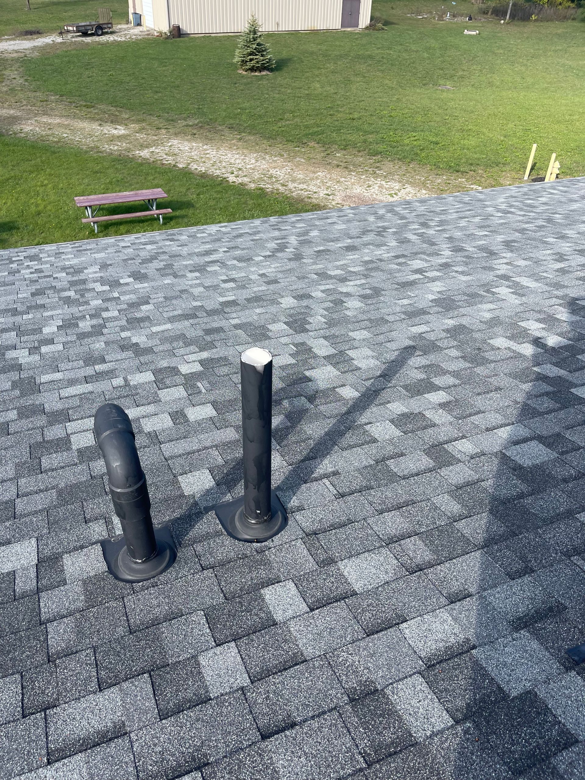 Rooftop with two black vent pipes and gray shingles, overlooking a grassy yard.