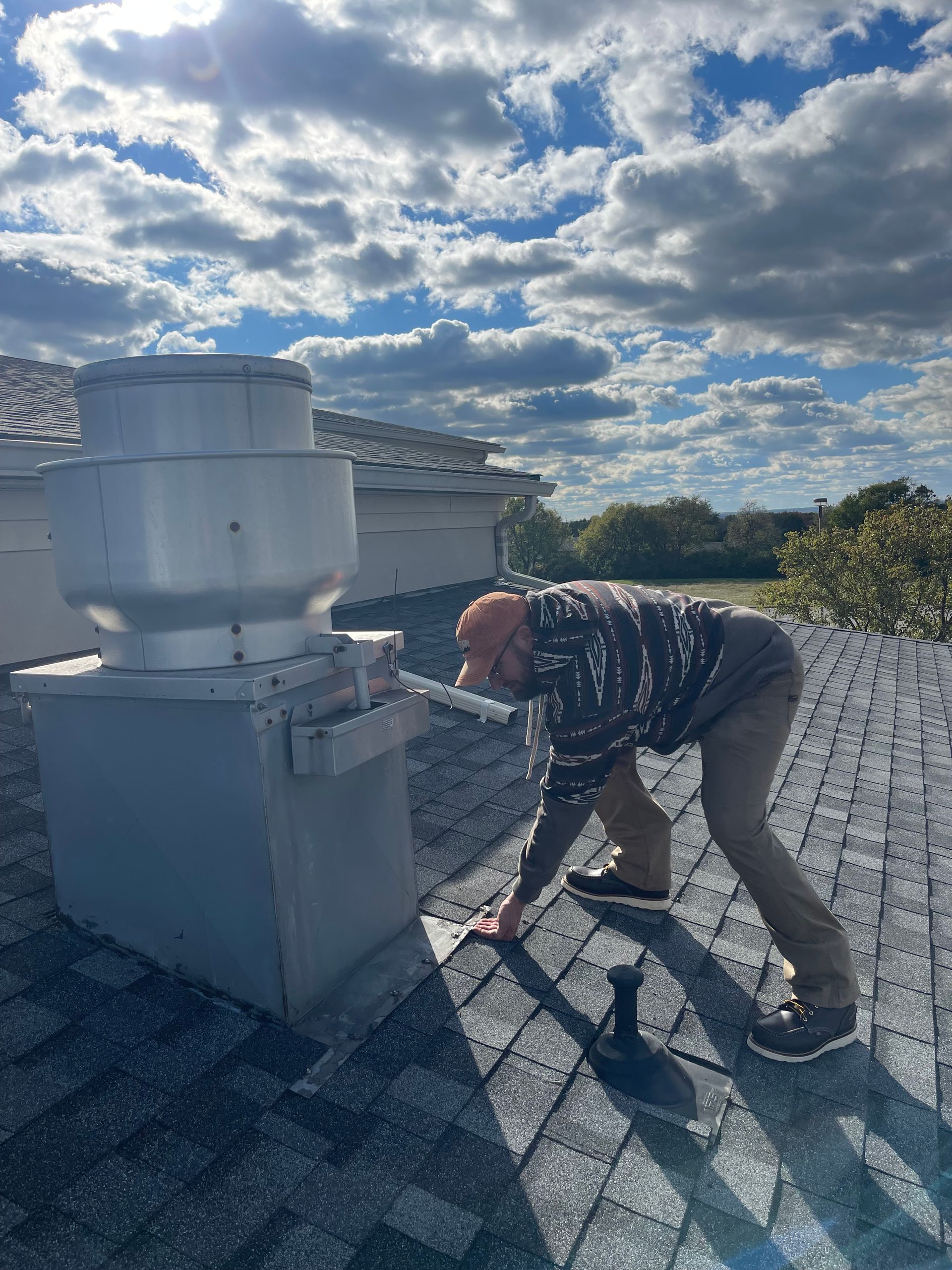 Worker repairing a rooftop vent on a shingled roof under a cloudy sky