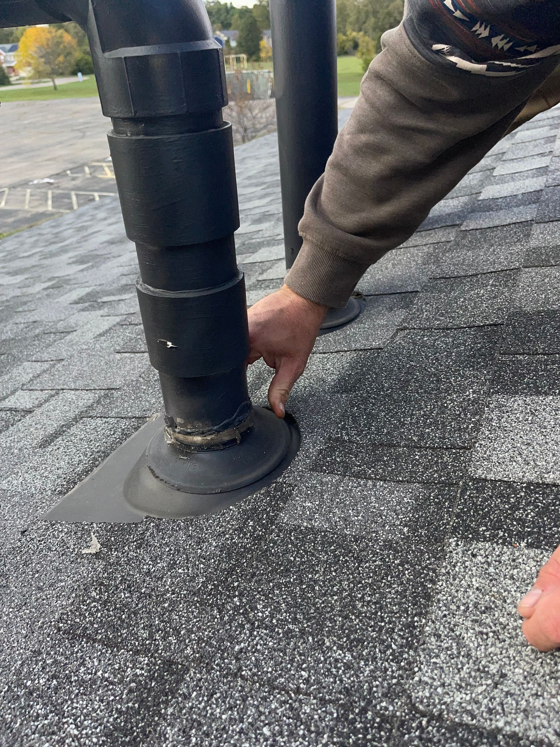 Worker sealing a black roof vent with roofing material on a gray shingled roof