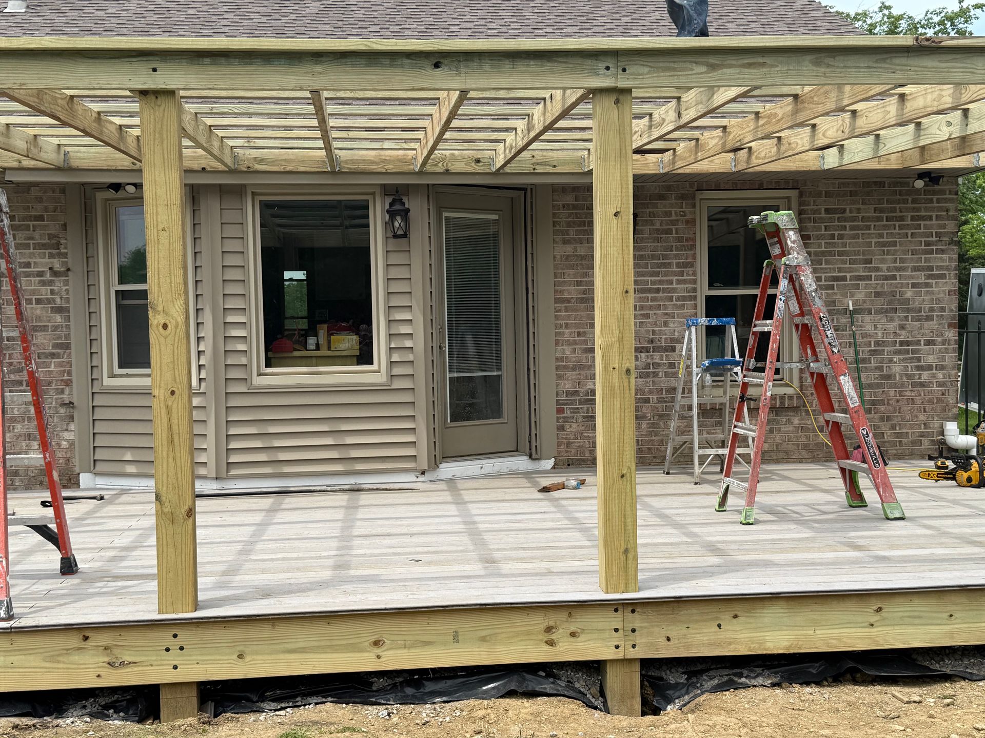 Partially built wooden deck and pergola in front of a house, with ladders and construction materials