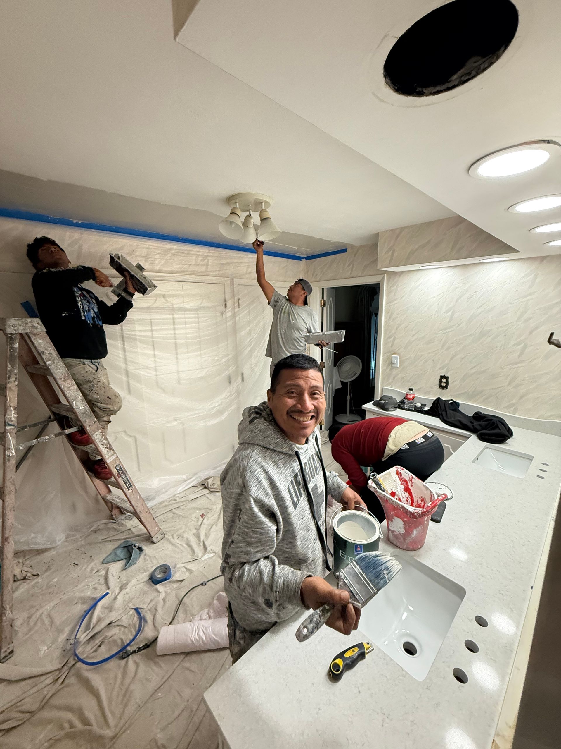 Workers painting a bathroom ceiling and walls, with one man smiling in the foreground holding a paint tray.