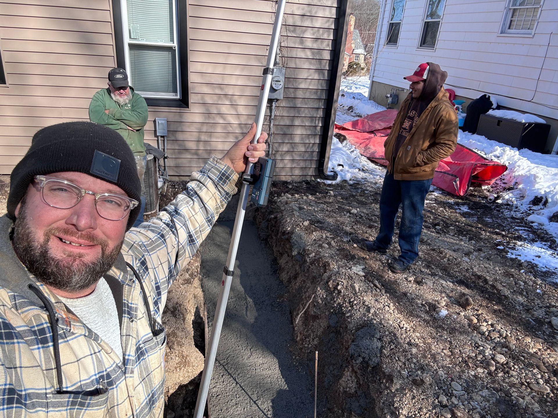 Three people outside a house beside a steep snowbank, one taking a selfie with a pole.