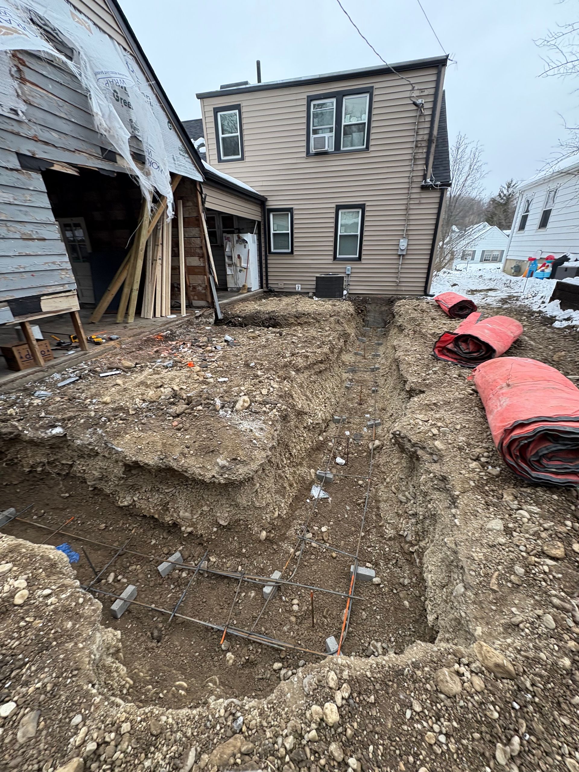Damaged house exterior with rubble and debris in a snowy yard, red pipes stacked on the right