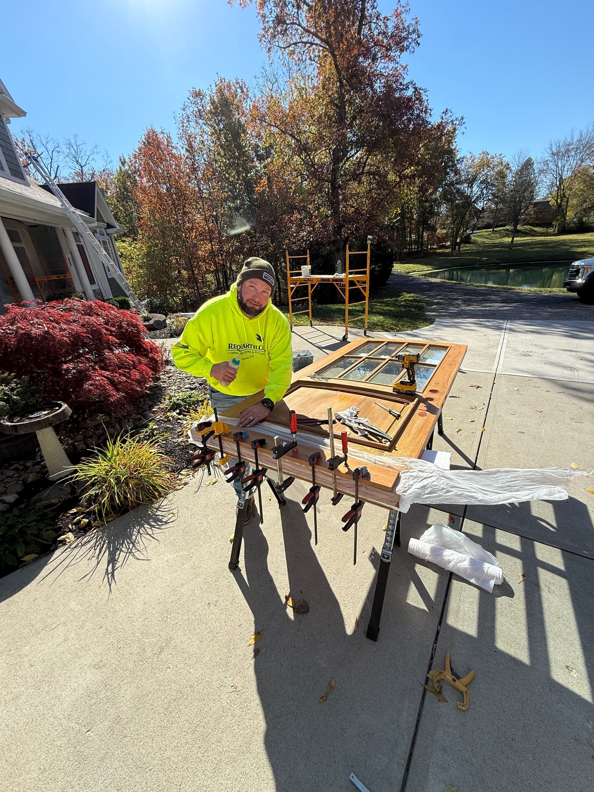 Worker in neon yellow shirt carrying a wooden frame in a sunny driveway with garden and house.