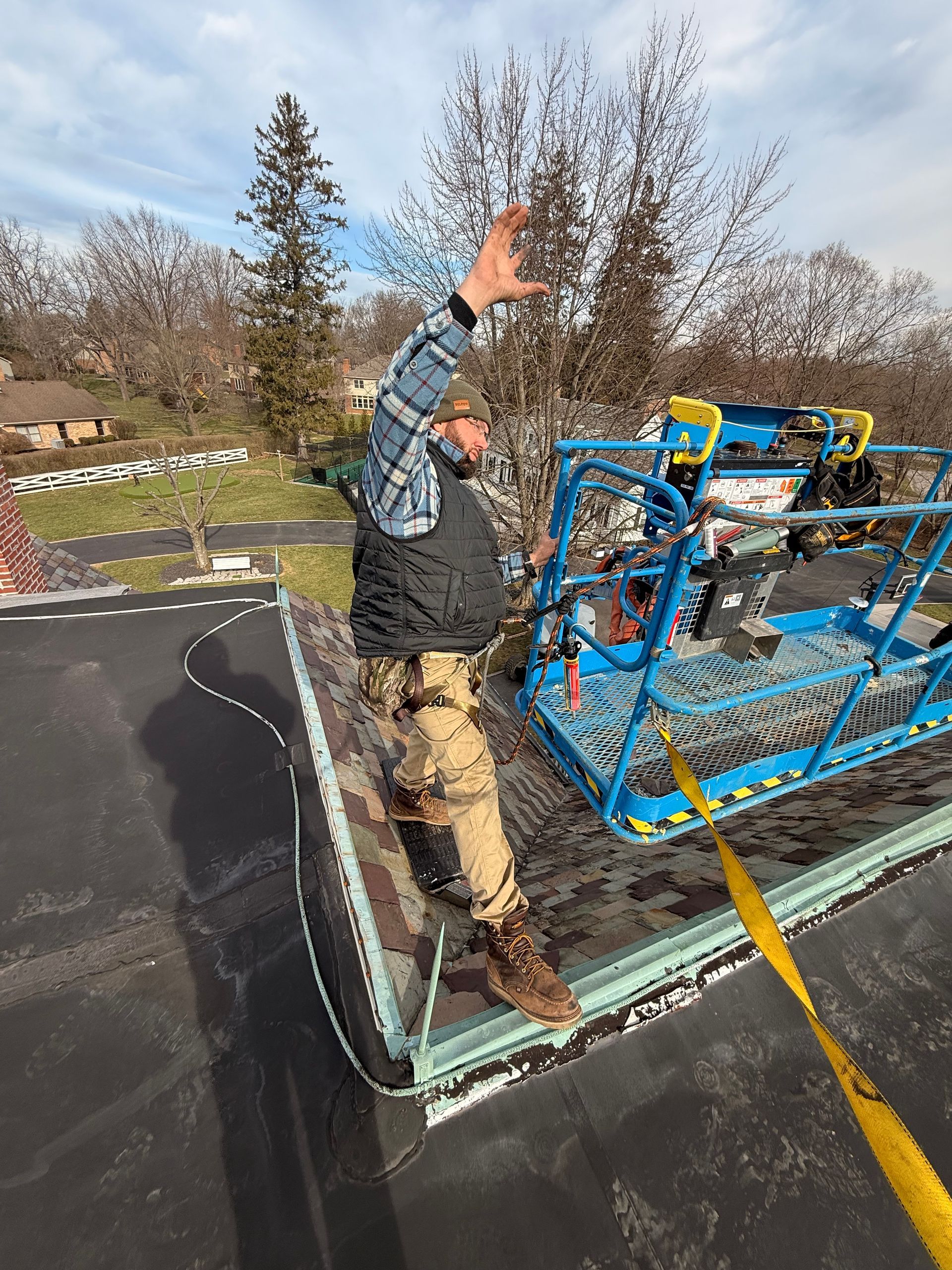 Worker standing on rooftop beside blue equipment, raising one arm near a yellow safety line.