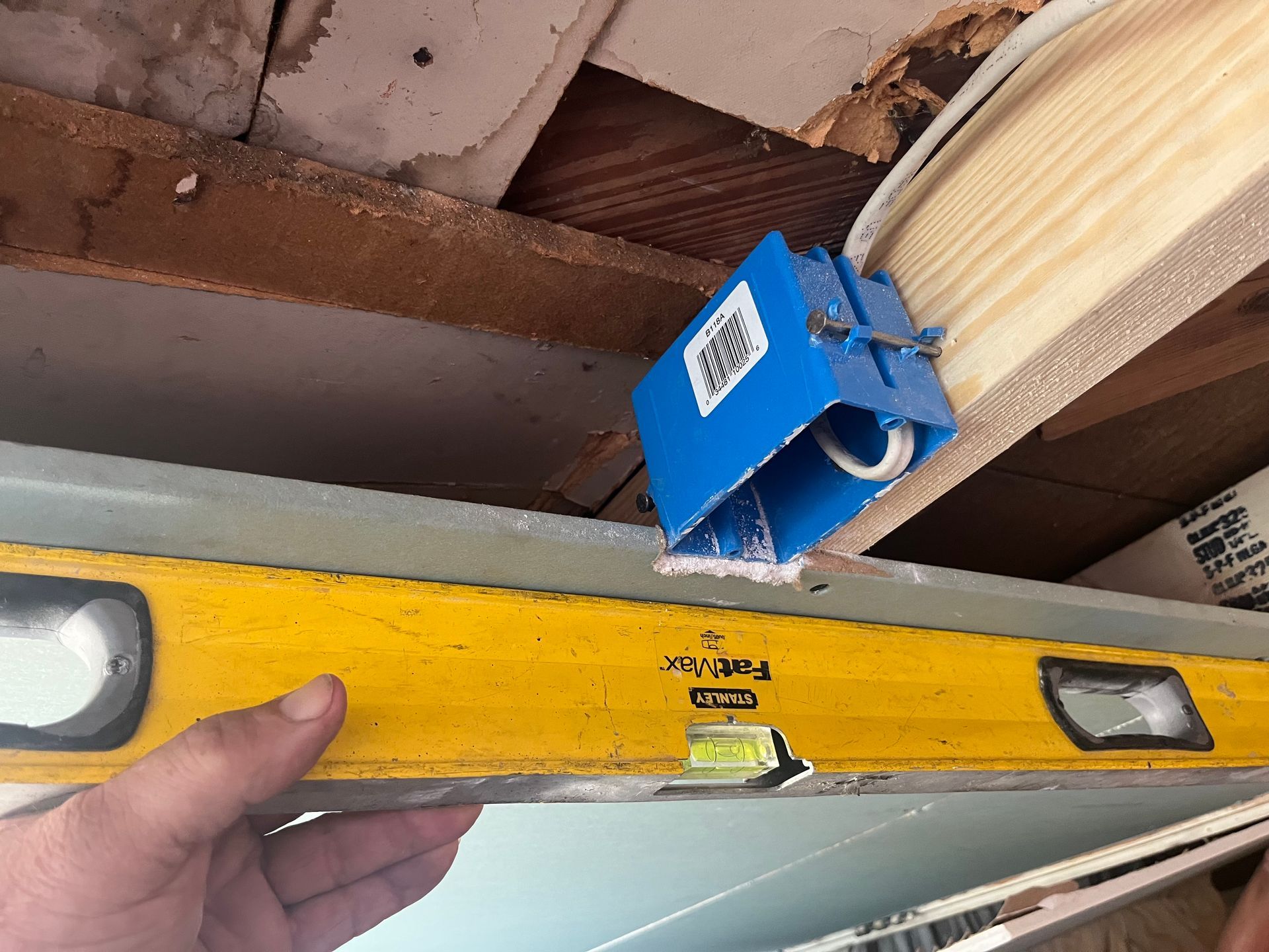 Hand holding a yellow level against a wooden beam beneath a blue electrical box in a ceiling area