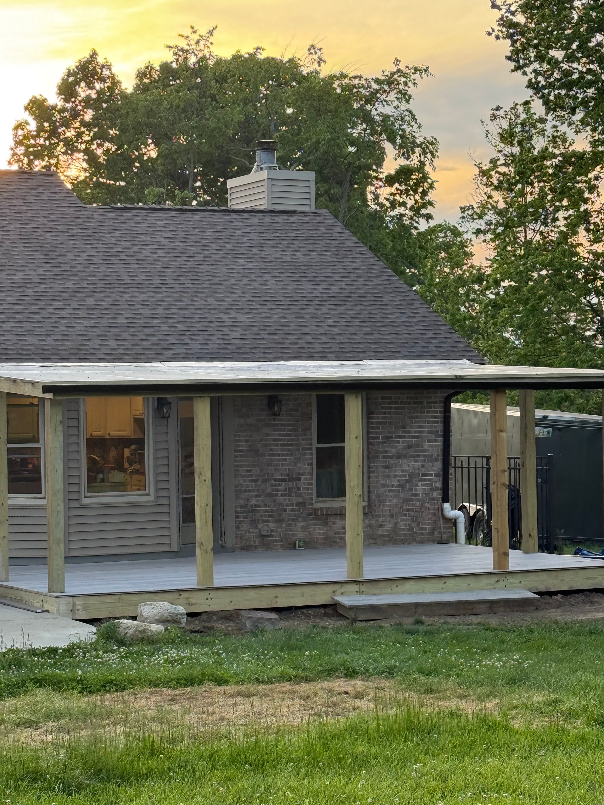 Small gray house with covered porch and chimney at sunset, framed by trees and grass.