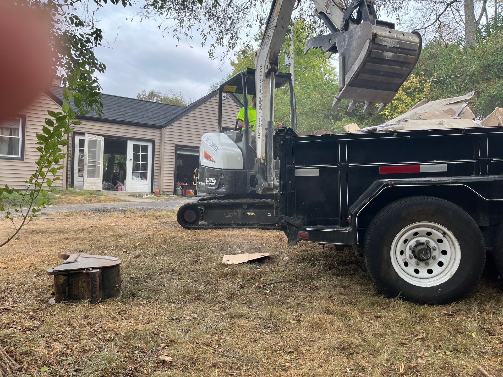 Excavator loading debris into a black dump truck in a residential yard outside a small house.