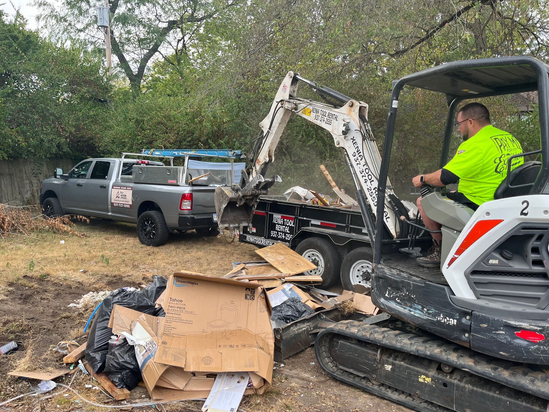 Excavator clearing debris beside damaged pickup trucks on a wooded roadside scene
