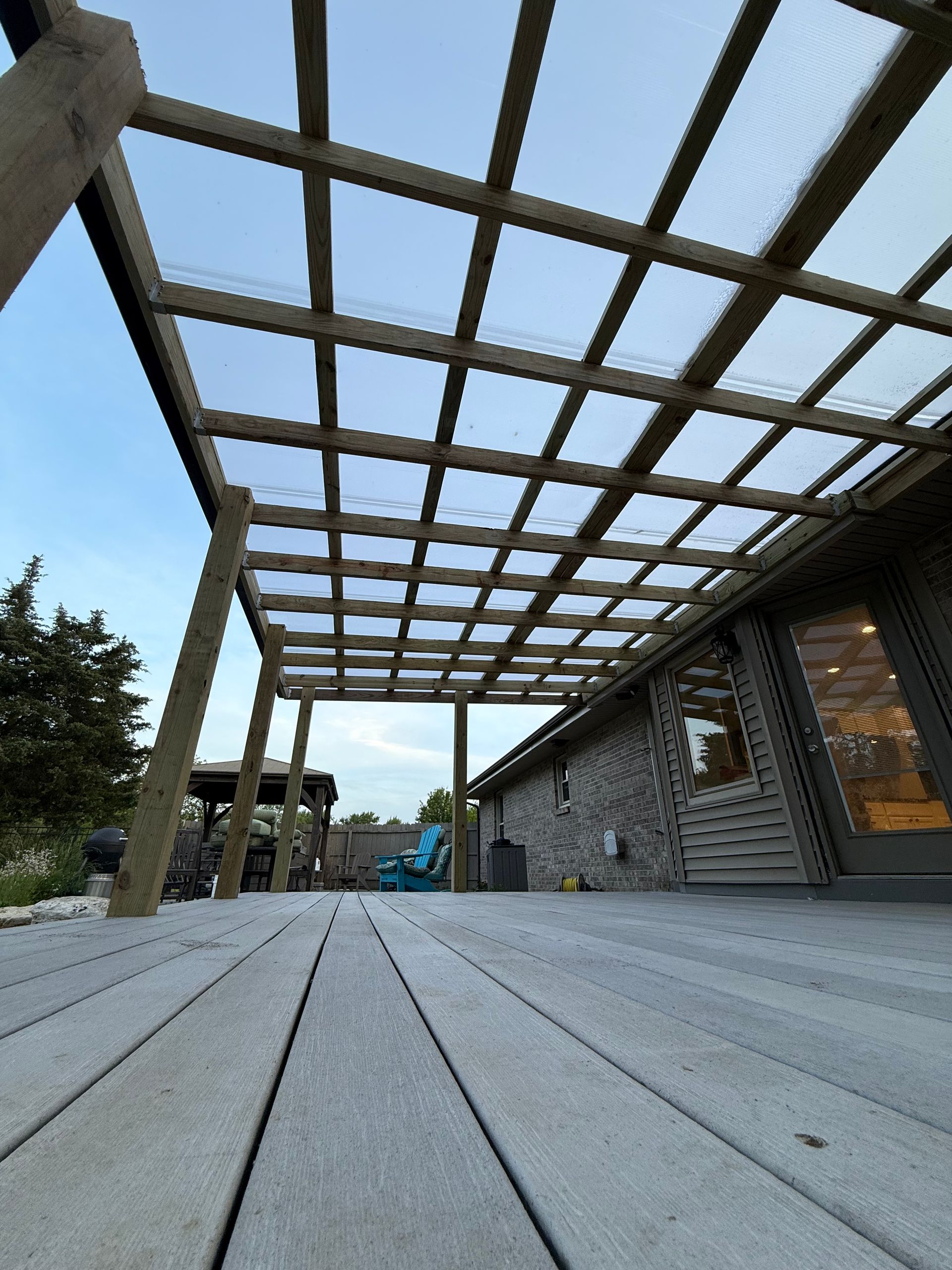 Low-angle view of a wooden deck beside a stone building under a slatted pergola structure.