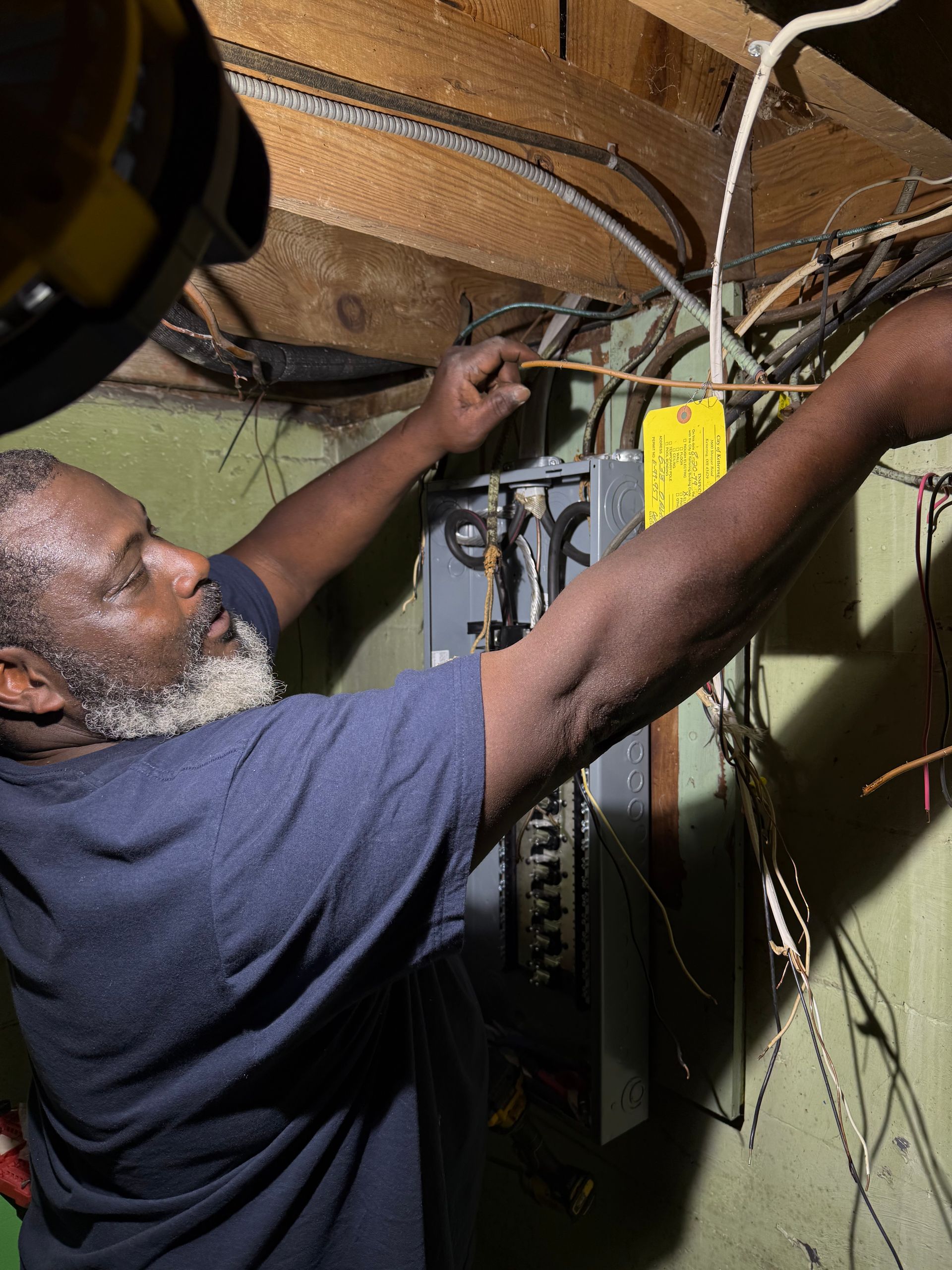 Technician wiring an electrical panel in a narrow utility space, reaching up under exposed cables.