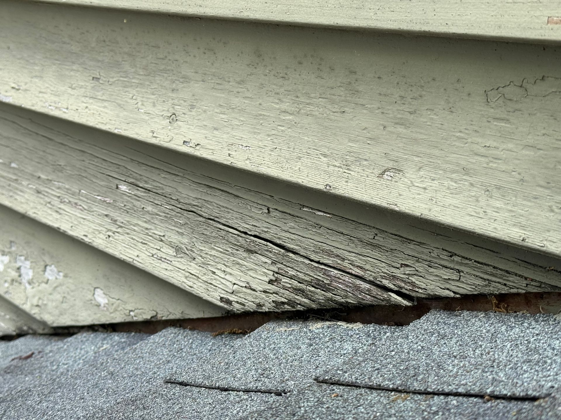 Close-up of damaged white vinyl siding with peeling paint above a gray shingled roof edge