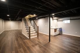 Finished basement with wood floors, exposed black ceiling, staircase, and small kitchenette.