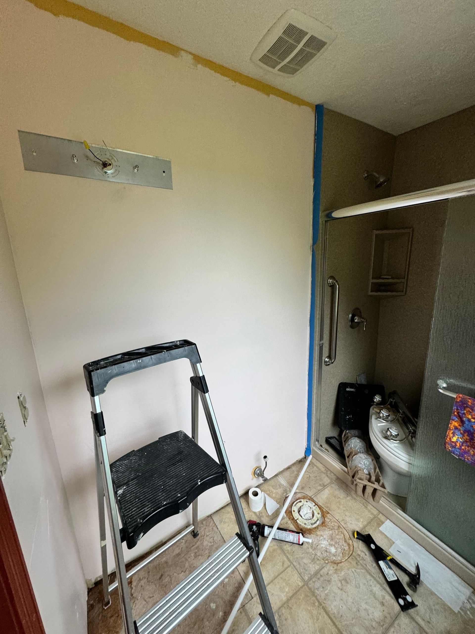 Small cluttered bathroom with a step stool, plumbing access panel, and cleaning supplies on the floor.
