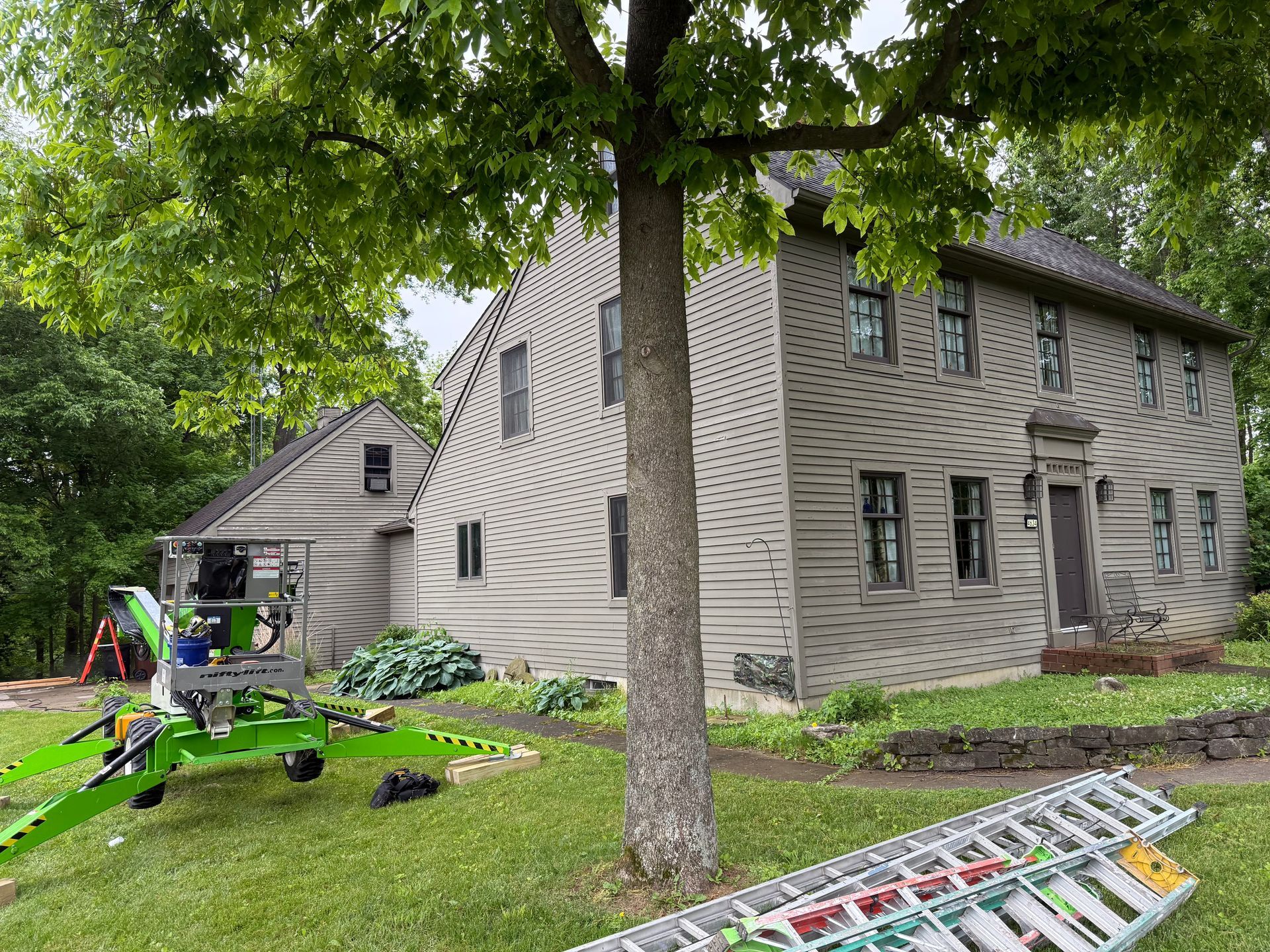 Ladder and tools on lawn beside a two-story house under trees, with workers near an extension