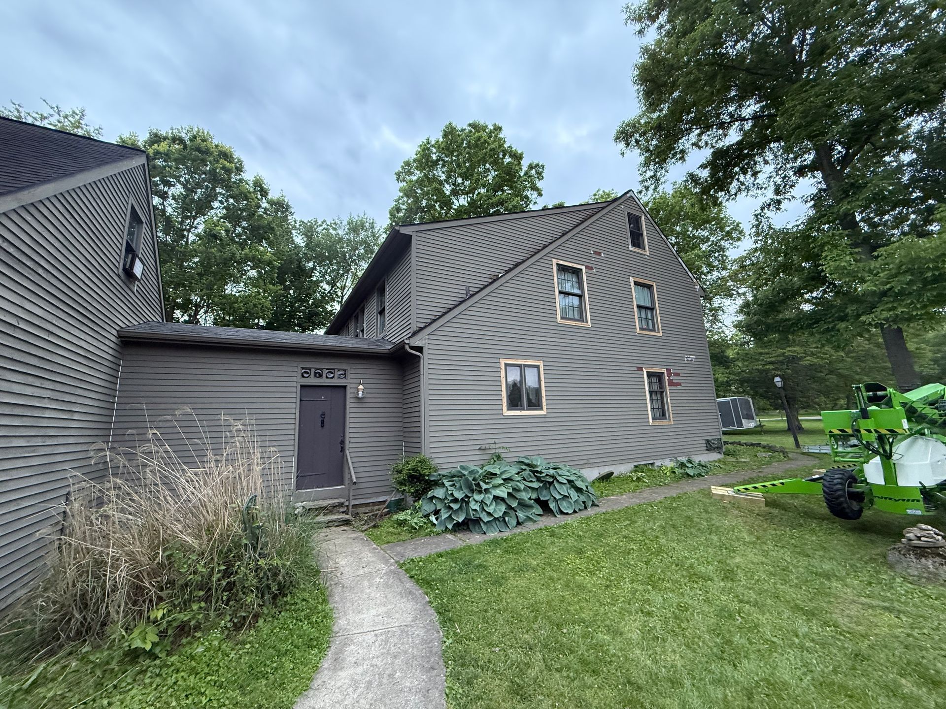 Gray house with steep roof, walkway, and green lawn beside a parked lawn tractor under trees