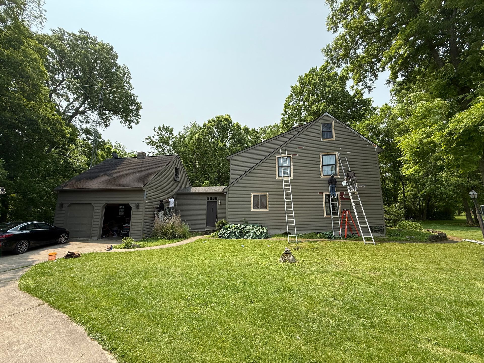 Person on ladder painting a gray house beside a detached garage in a green yard