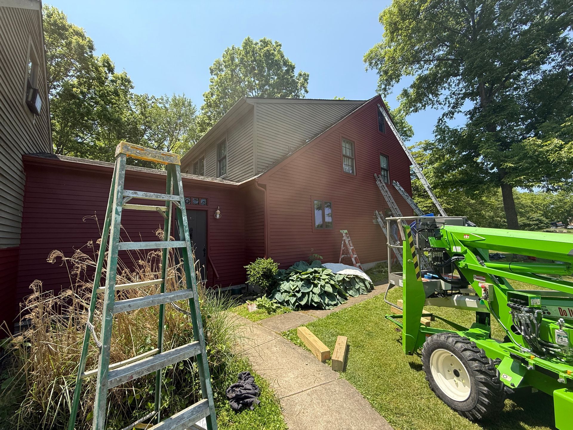 Ladder leaning against red house; green tractor parked on a grassy path in a sunny yard.