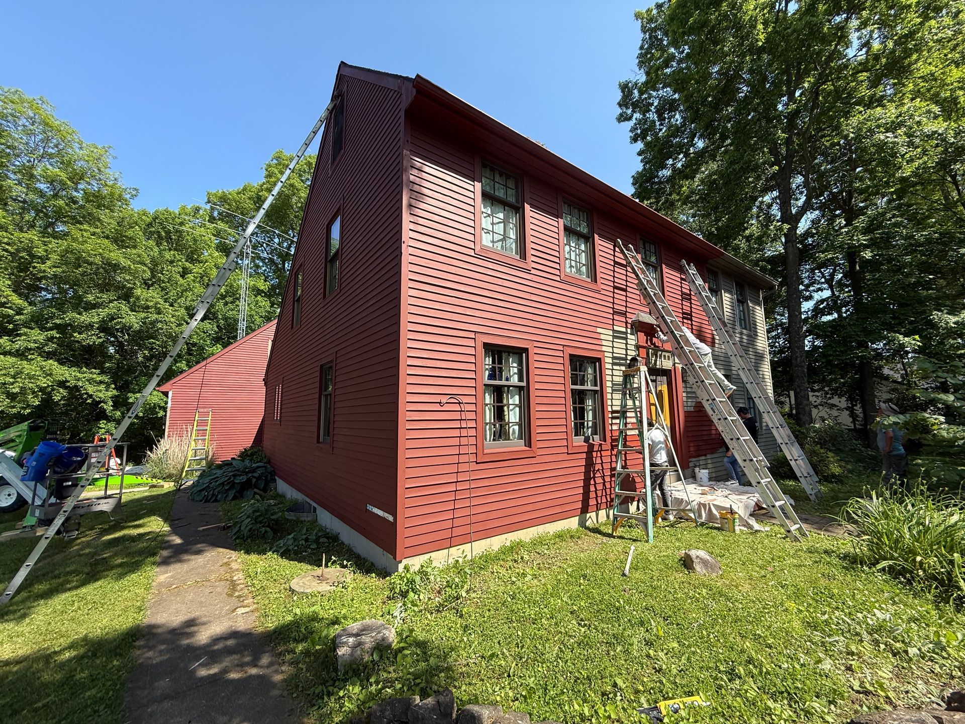 Red two-story house under renovation with ladders on the side, surrounded by trees and grass