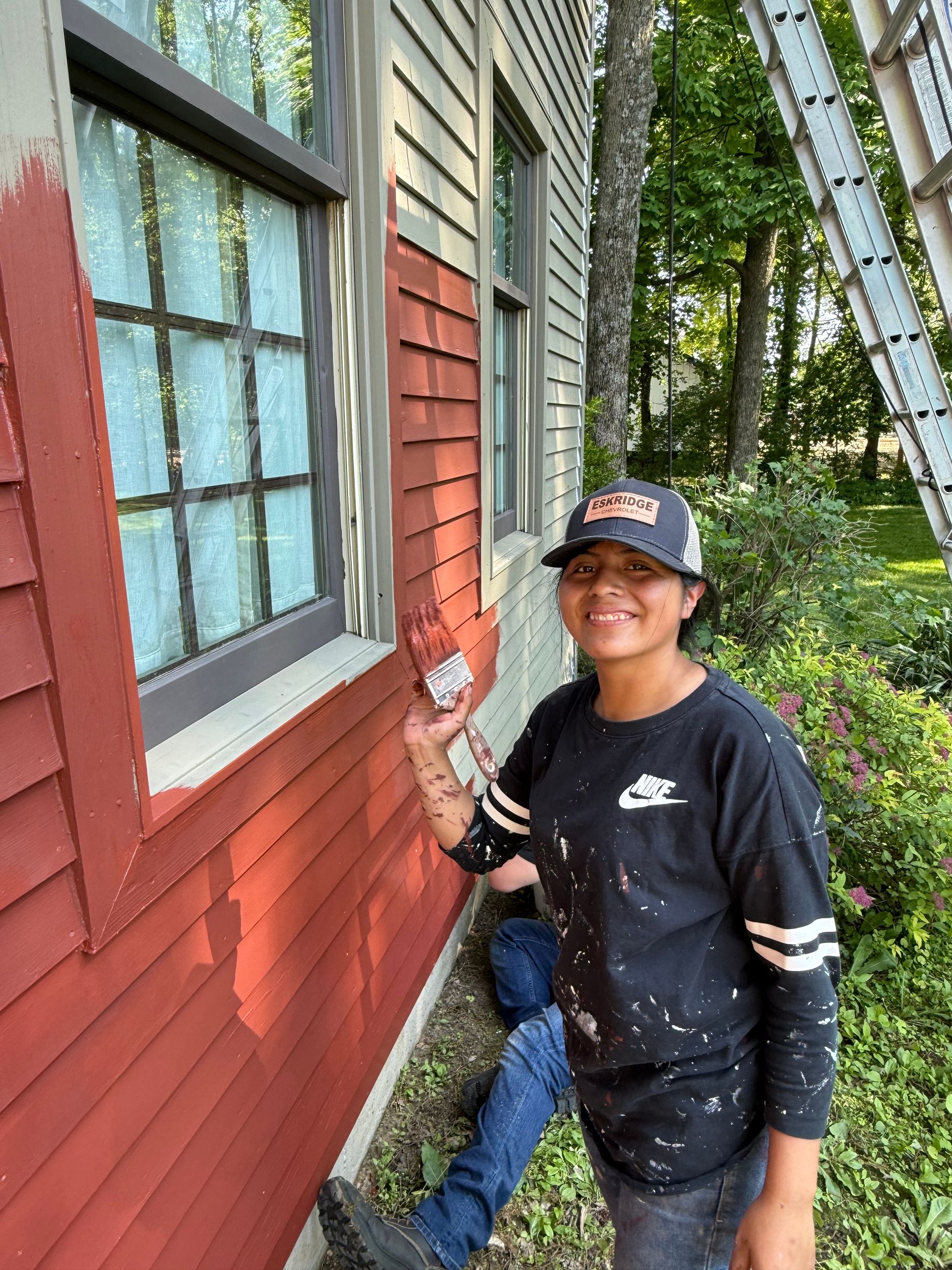 Person painting a red house exterior near a window, smiling and holding a paintbrush.