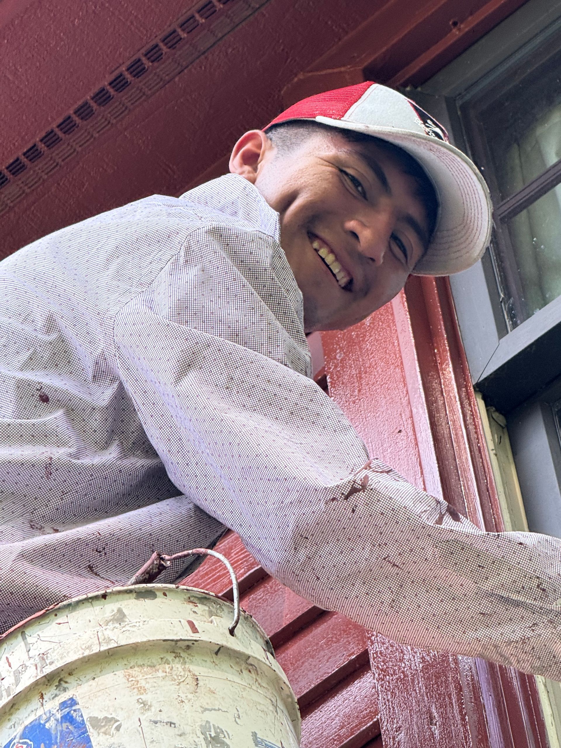 Person smiling while painting a red wall, wearing a white cap and carrying a paint bucket.