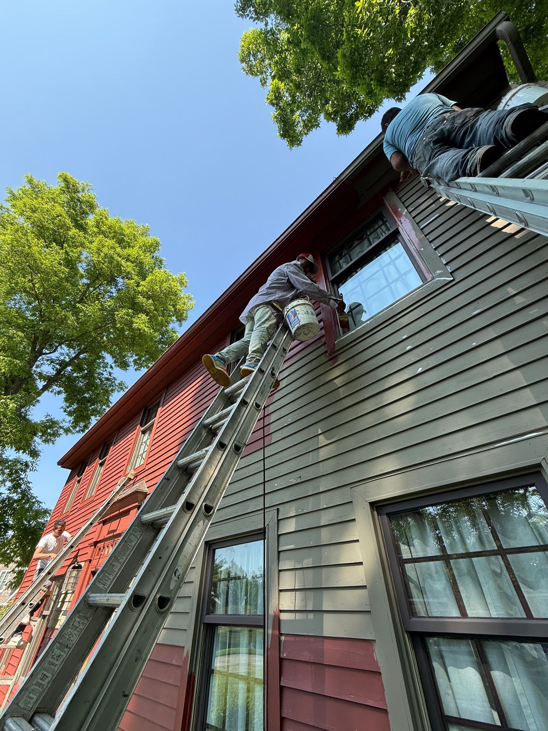 Worker on ladder repairing red house roof beside leafy trees