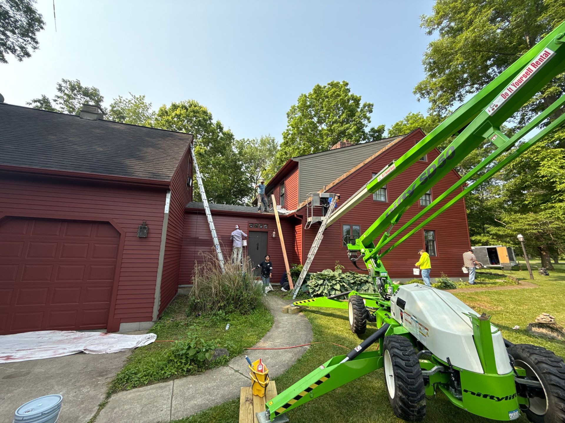 Bright green lift beside red house; worker on ladder repairs roof amid scattered debris.