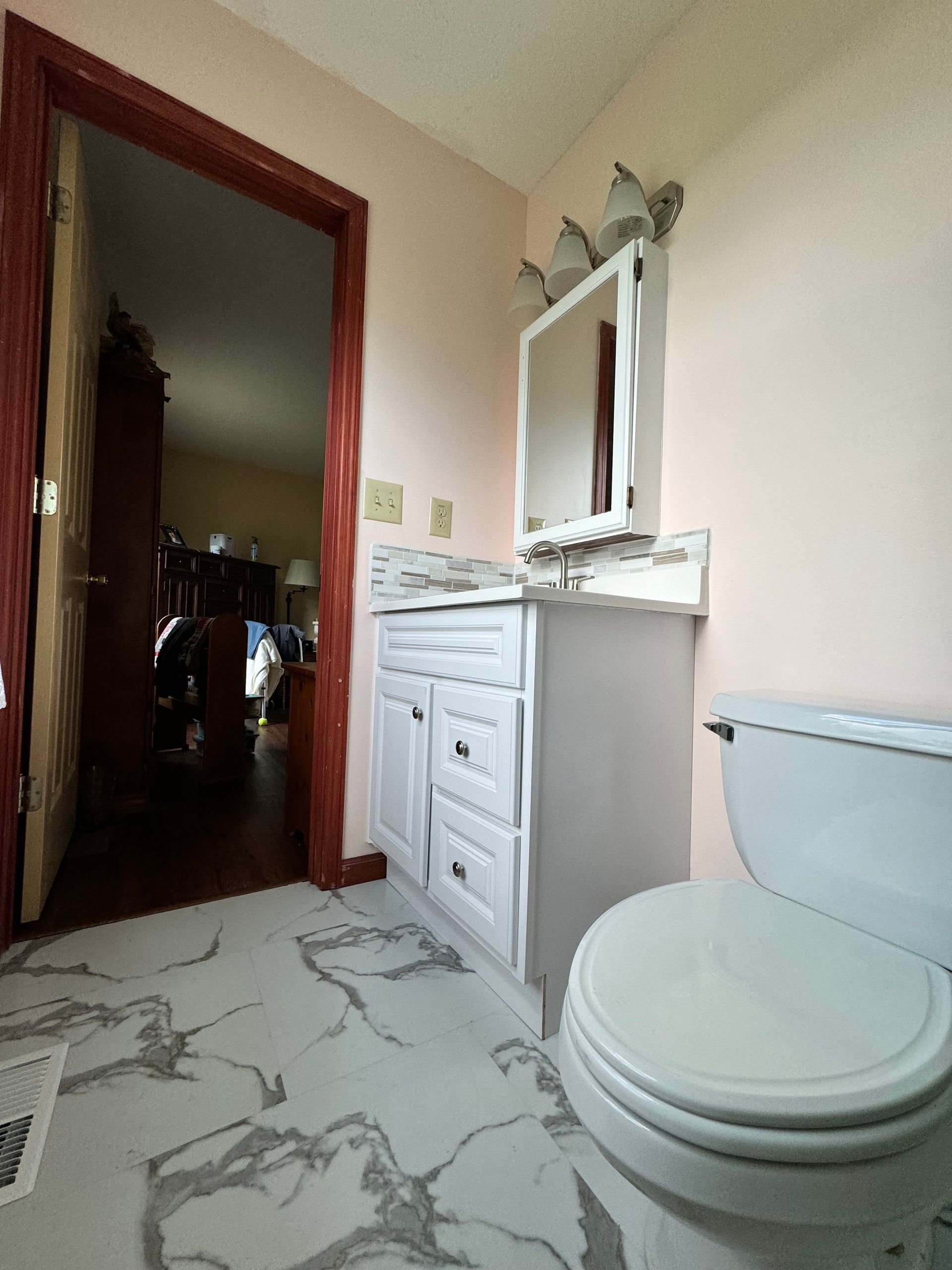 Small bathroom with white toilet, vanity, mirror, and beige walls; doorway opens to a darker room.