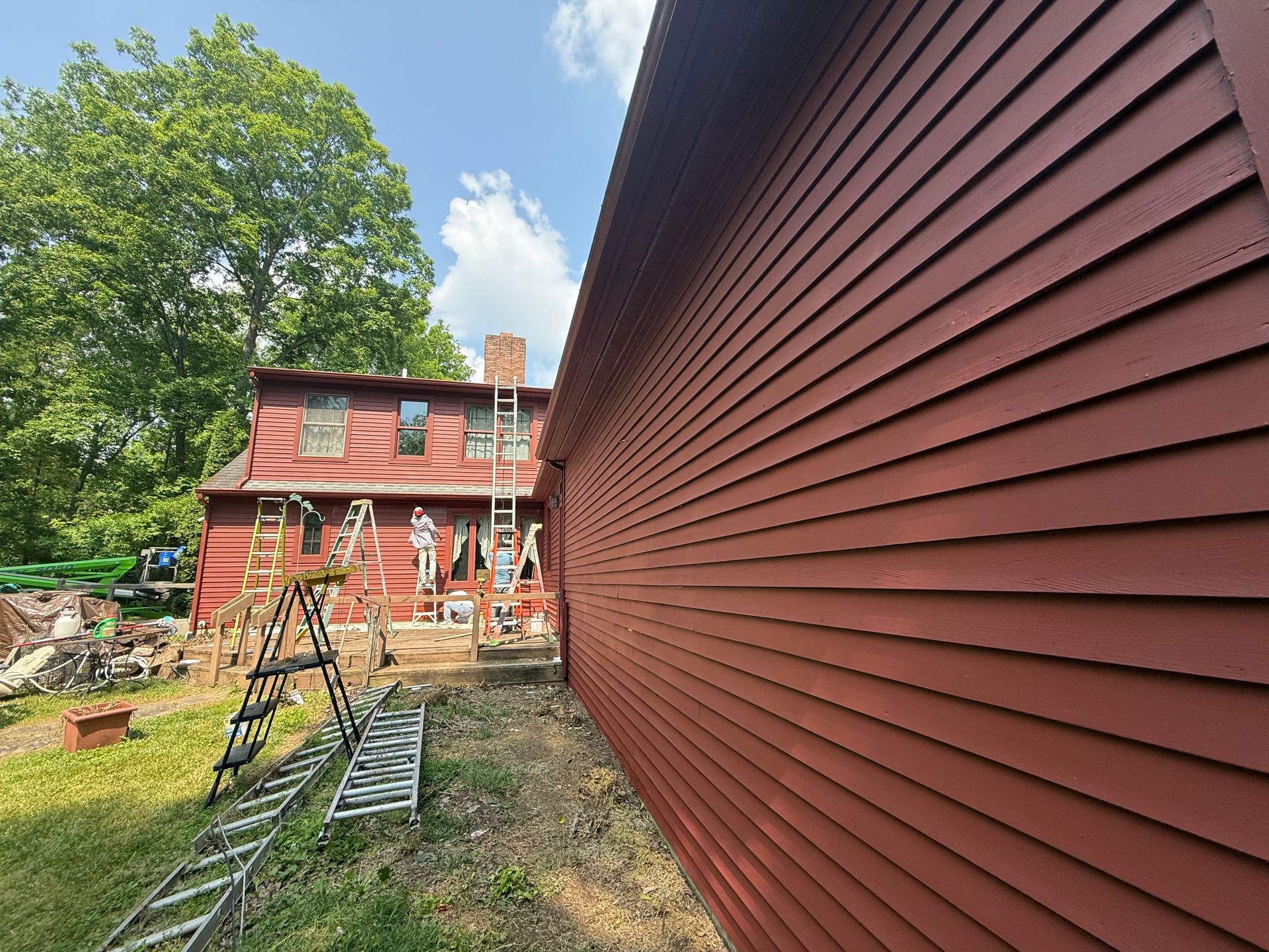 Red house siding dominates foreground; another red house with scaffolding and ladder stands across a grassy yard.