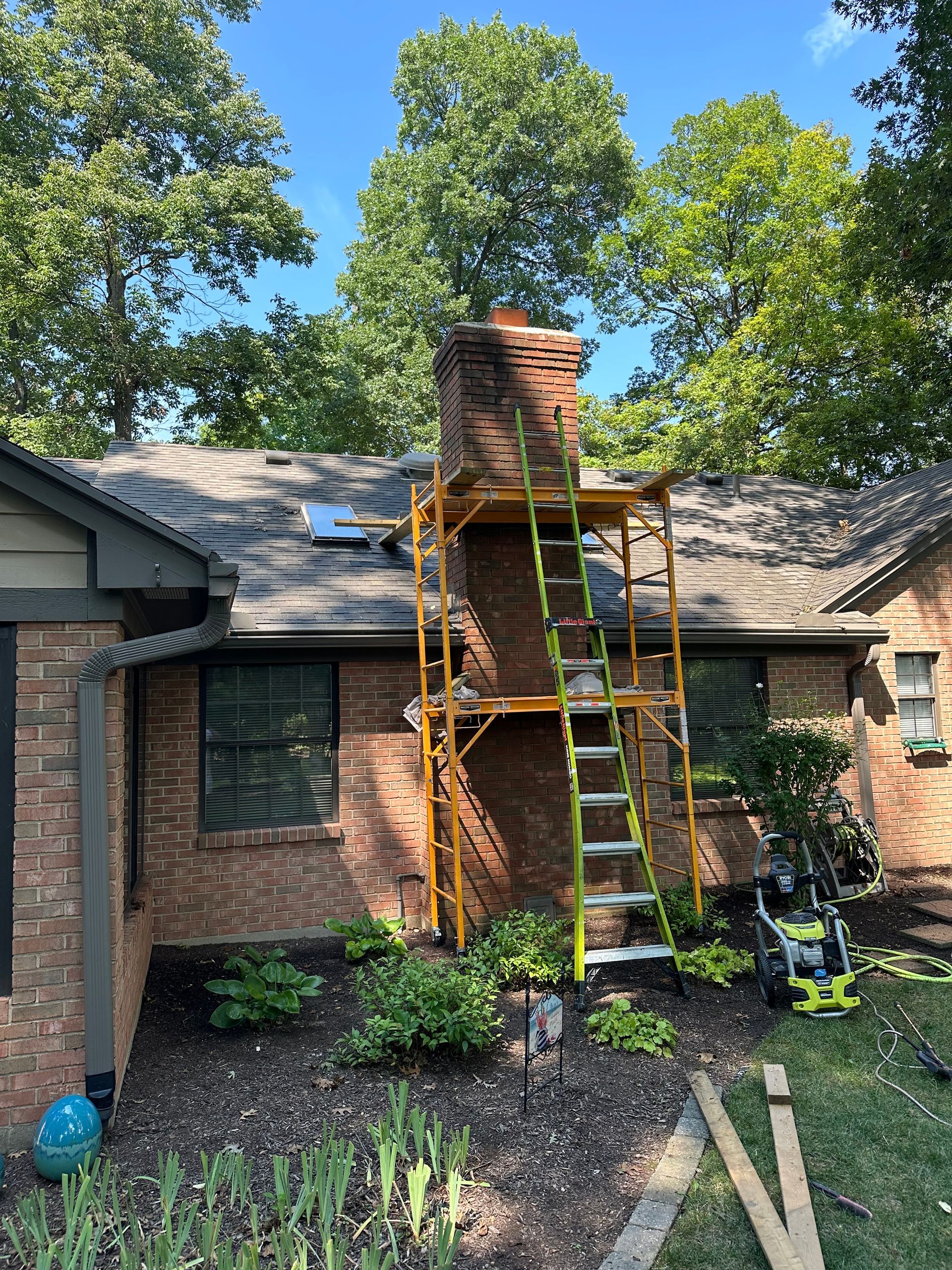 Worker repairing brick chimney on a house roof with scaffolding and ladder, trees in background