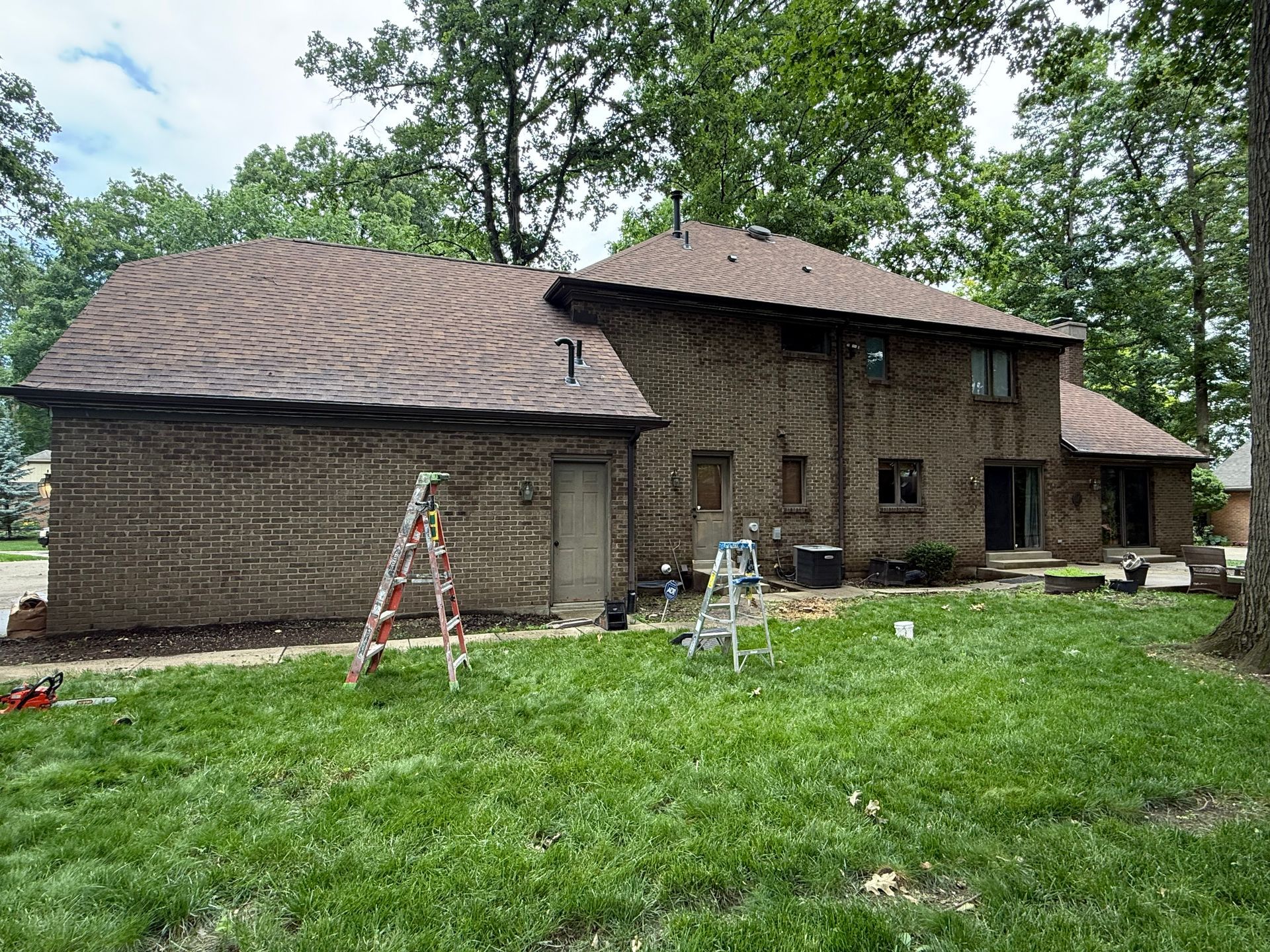 Two-story brick house with ladders in the yard, surrounded by trees and grass