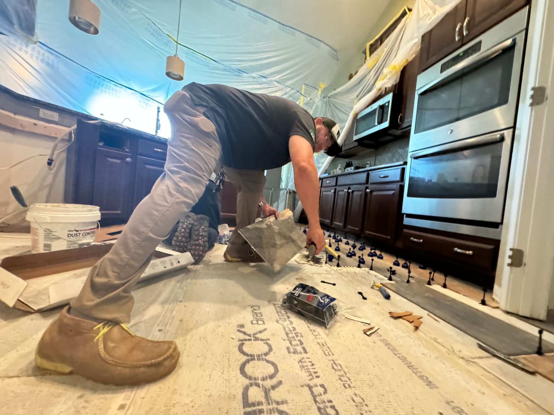 Worker installing flooring in a kitchen, bending over with tools near cabinets and built-in ovens.