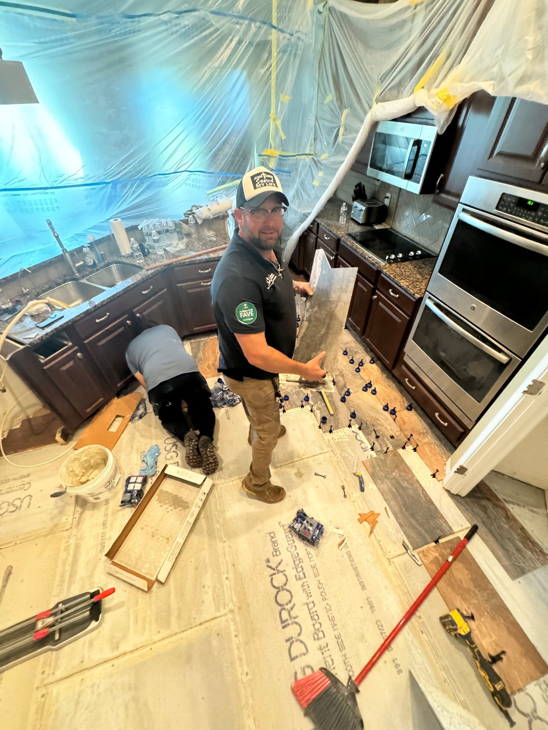 Worker crouches in a kitchen renovation, surrounded by tools and debris, with cabinets and appliances along the wall.