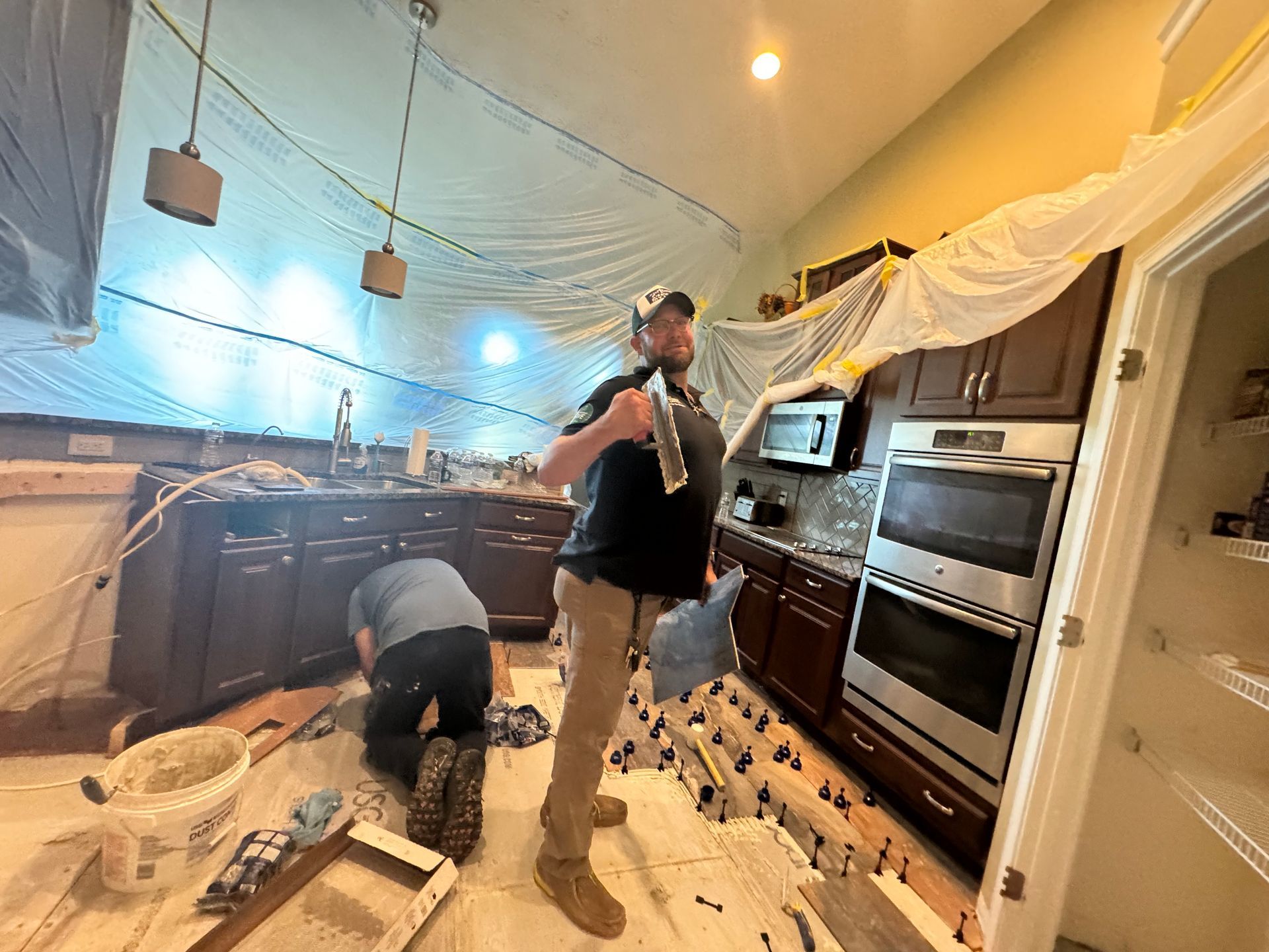 Two workers installing a kitchen with cabinets, appliances, and string lights under a draped ceiling.