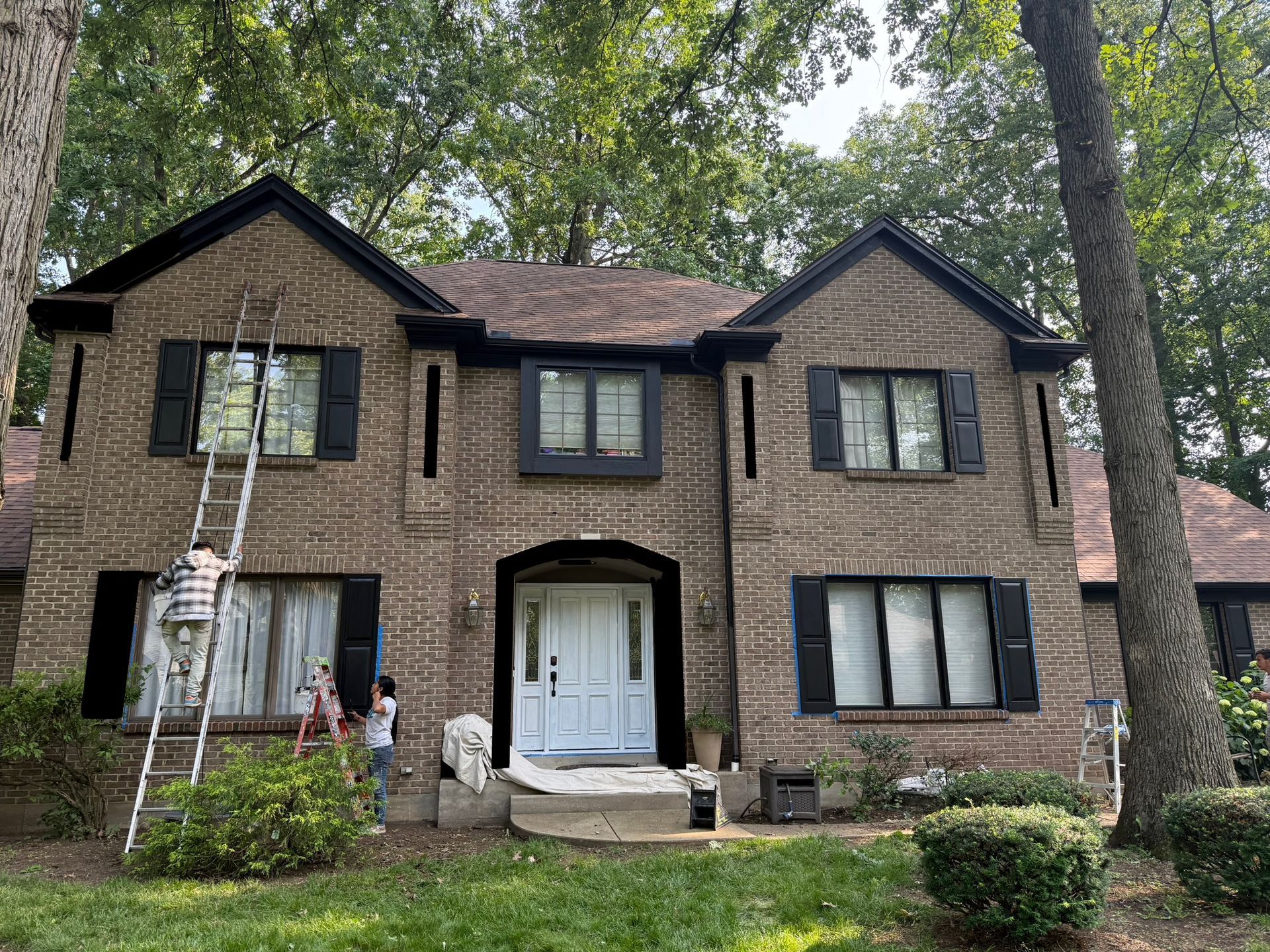 Two-story brick house with white front doors and a ladder leaning against the left side, surrounded by trees