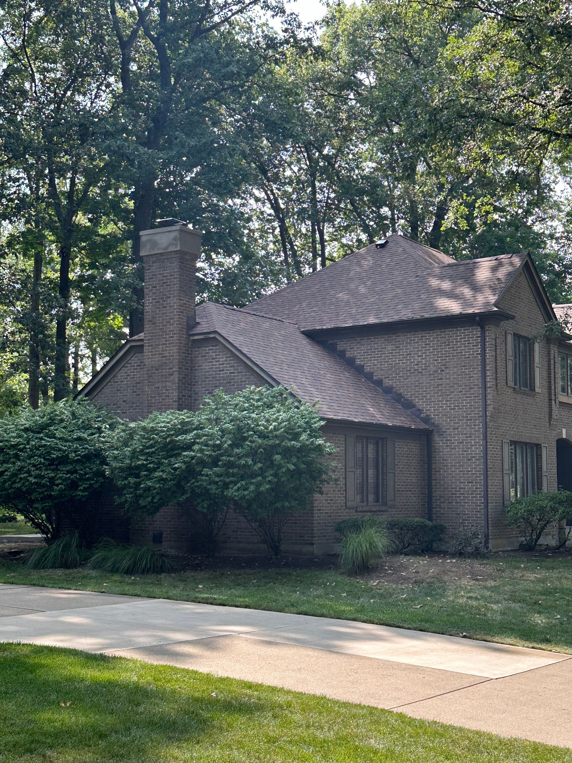 Brick house with a chimney and wavy roof, partly shaded by trees along a sidewalk
