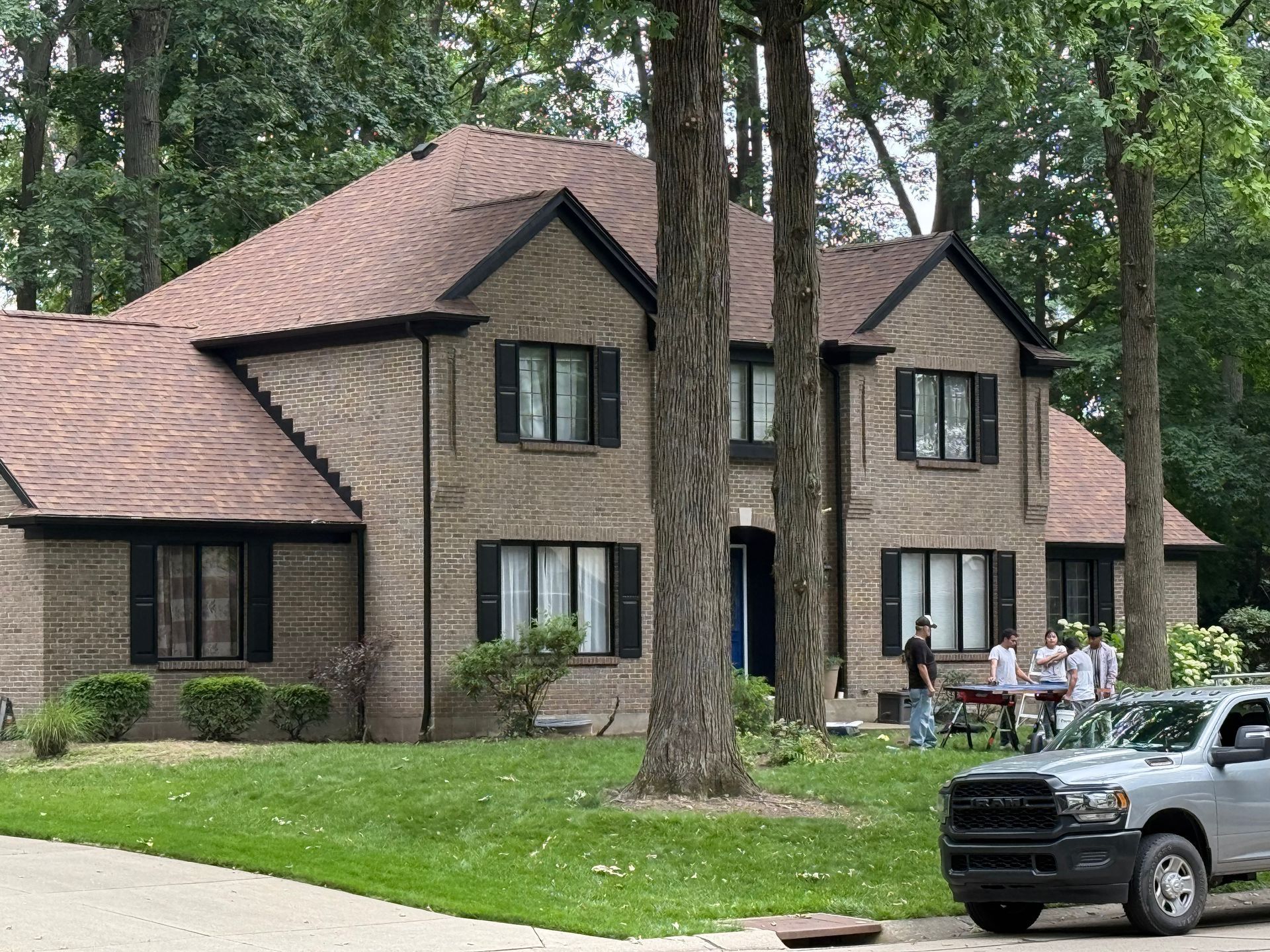 Two-story tan house with brown roof in a wooded yard, with a pickup truck and people near the front drive.