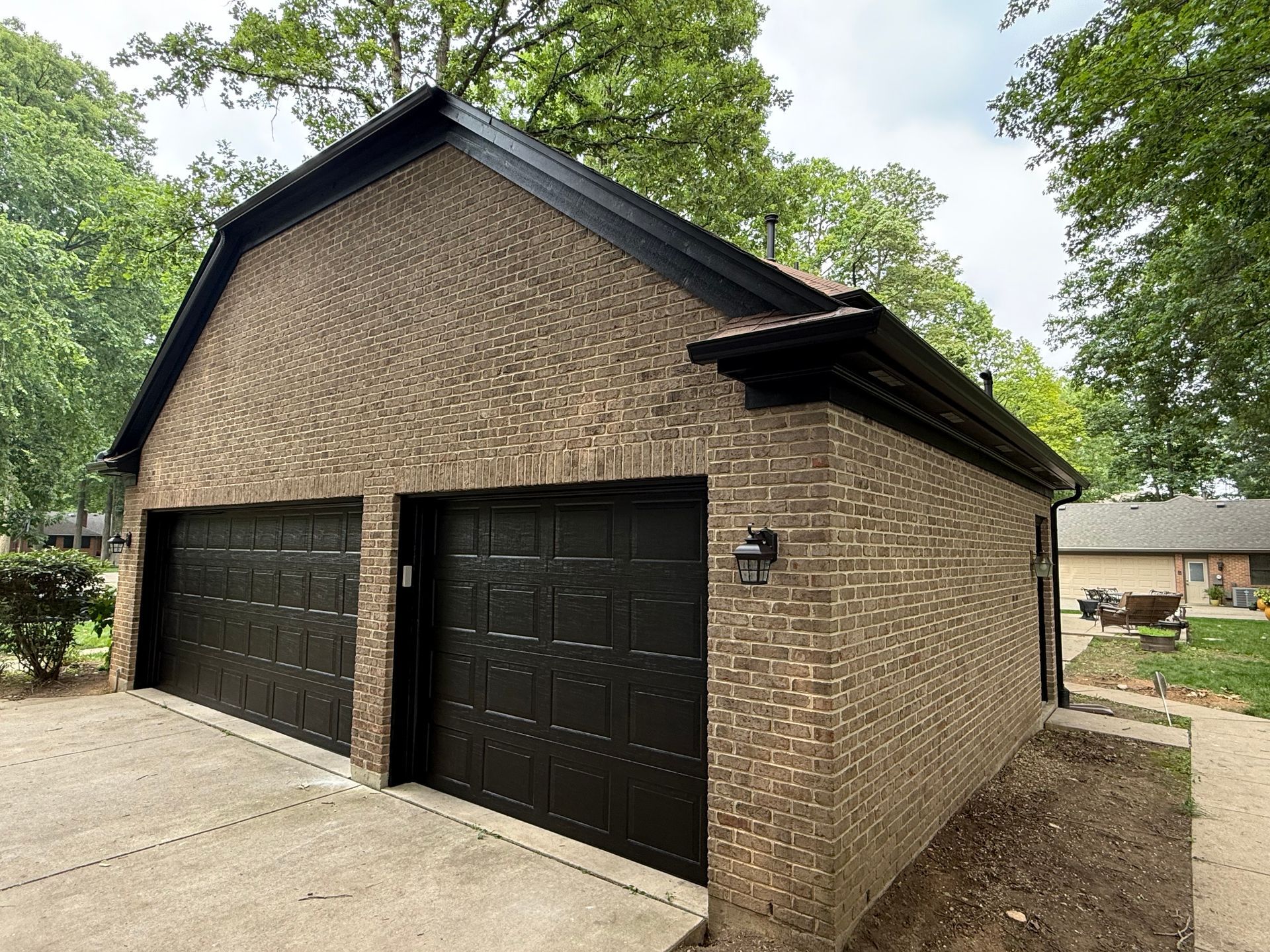 Brick two-car garage with black doors and a sloped roof, set beside a driveway and trees.