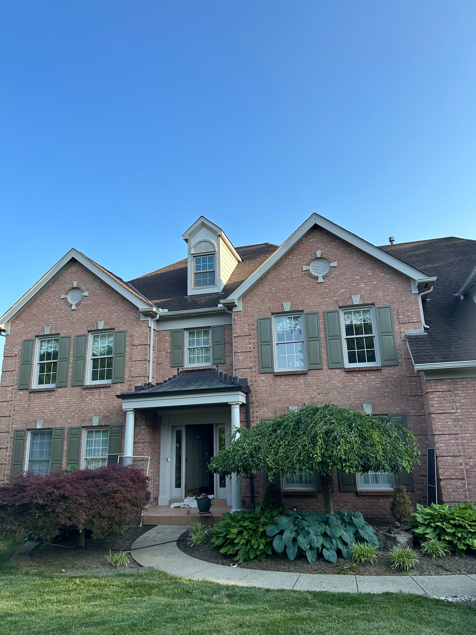 Brick suburban house with green shutters, arched entry, and manicured front landscaping under blue sky