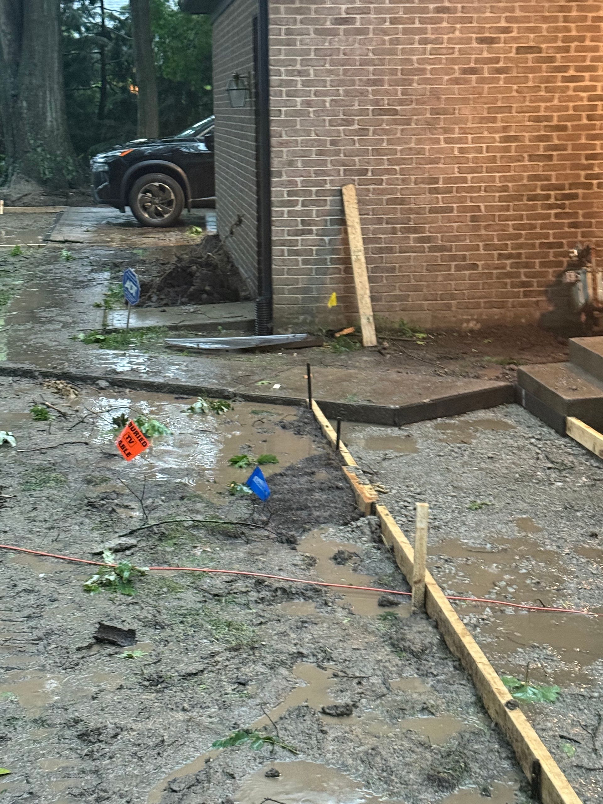 Construction site with muddy ground, wooden stakes, and a brick wall beside a parked vehicle.