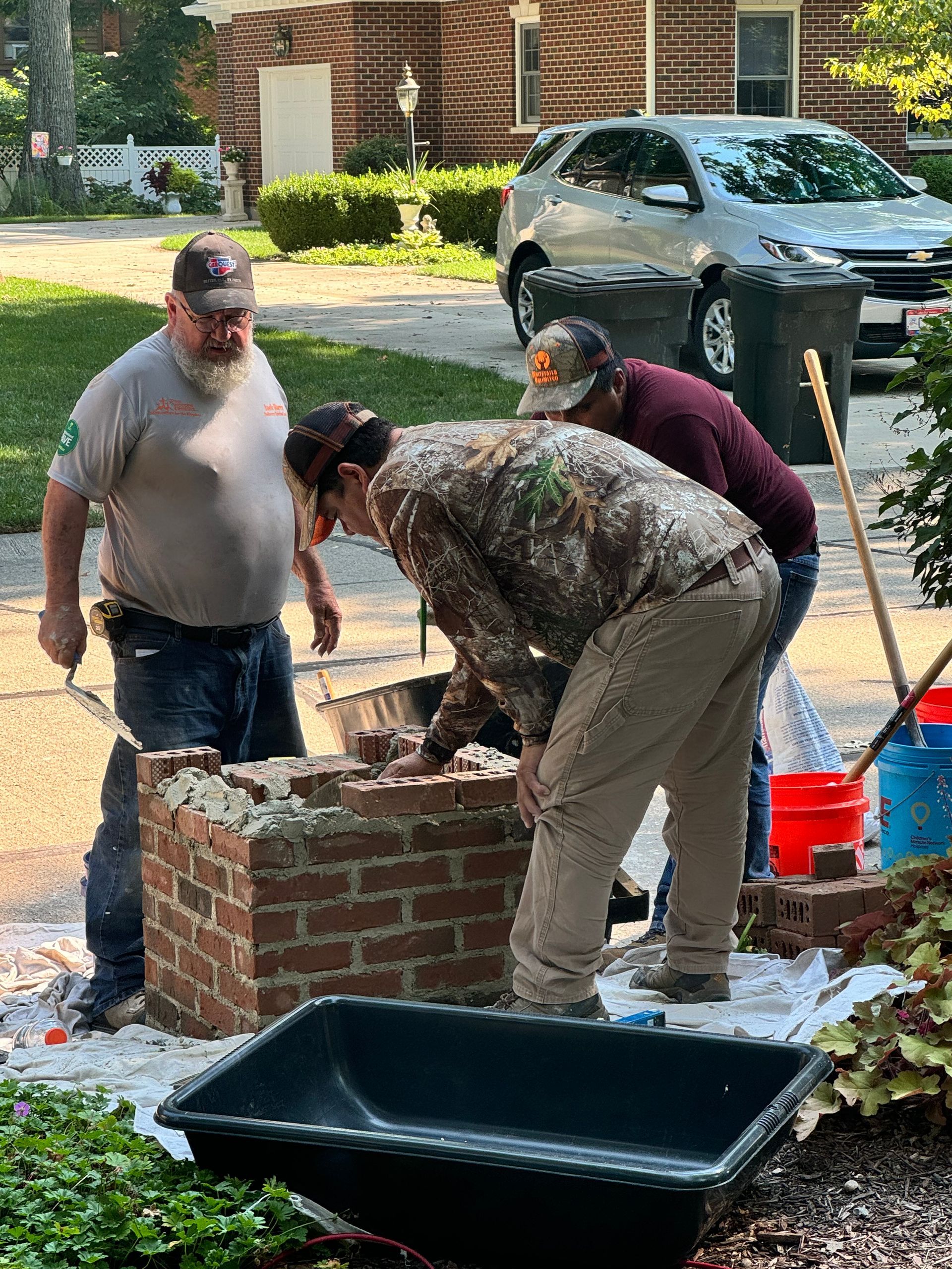 Workers building a brick planter beside a driveway, with tools and a car nearby.