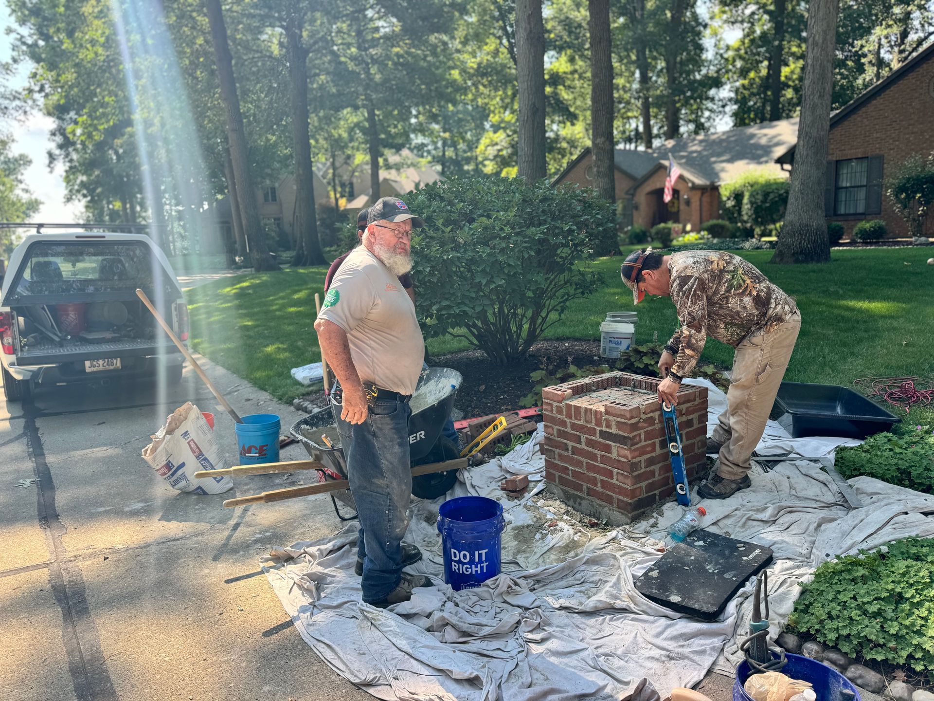 Two men working outdoors on a brick fire pit, with tools and supplies on drop cloths in a driveway