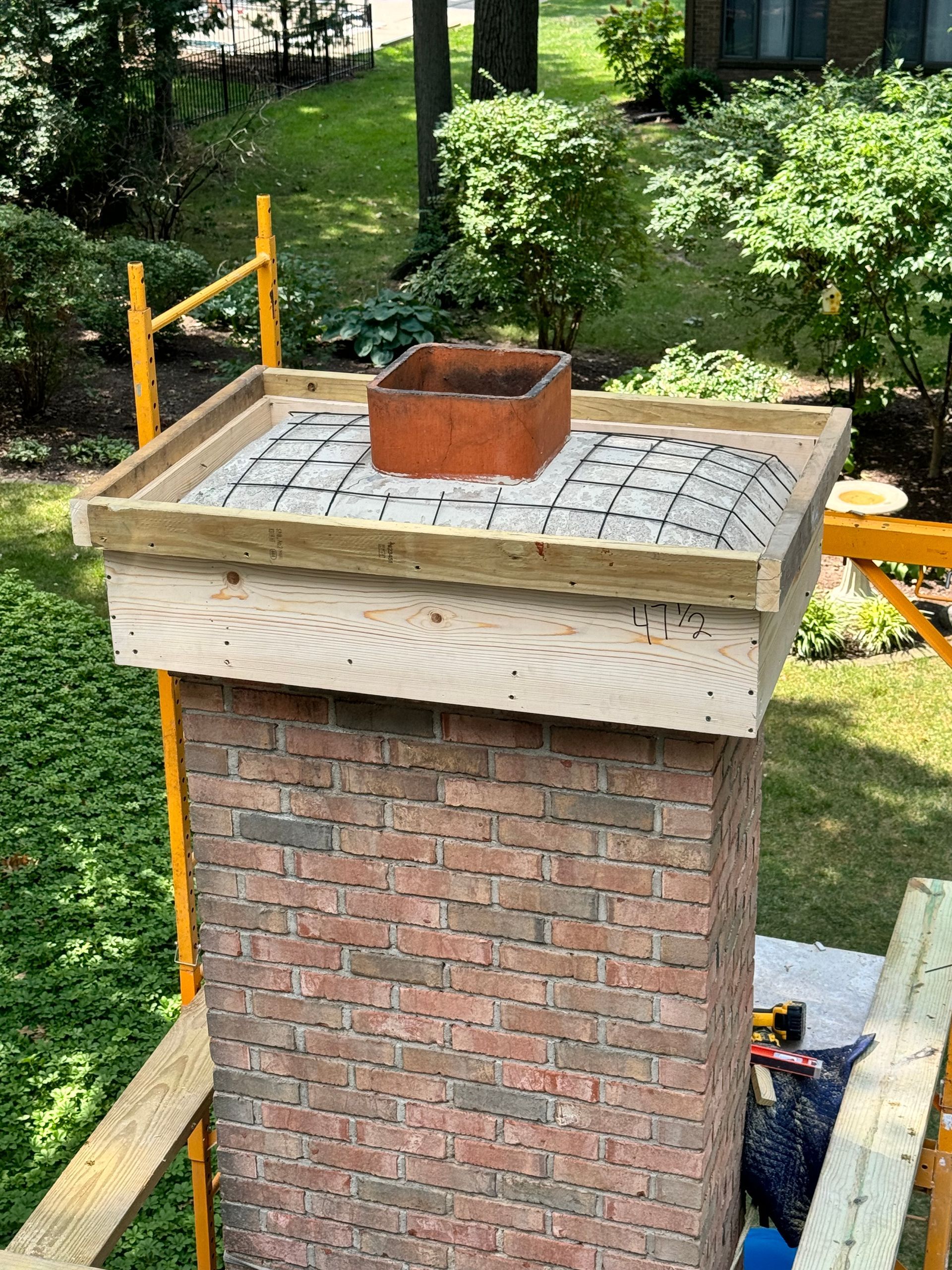 Brick chimney under construction with a metal flue opening on top, surrounded by scaffolding.