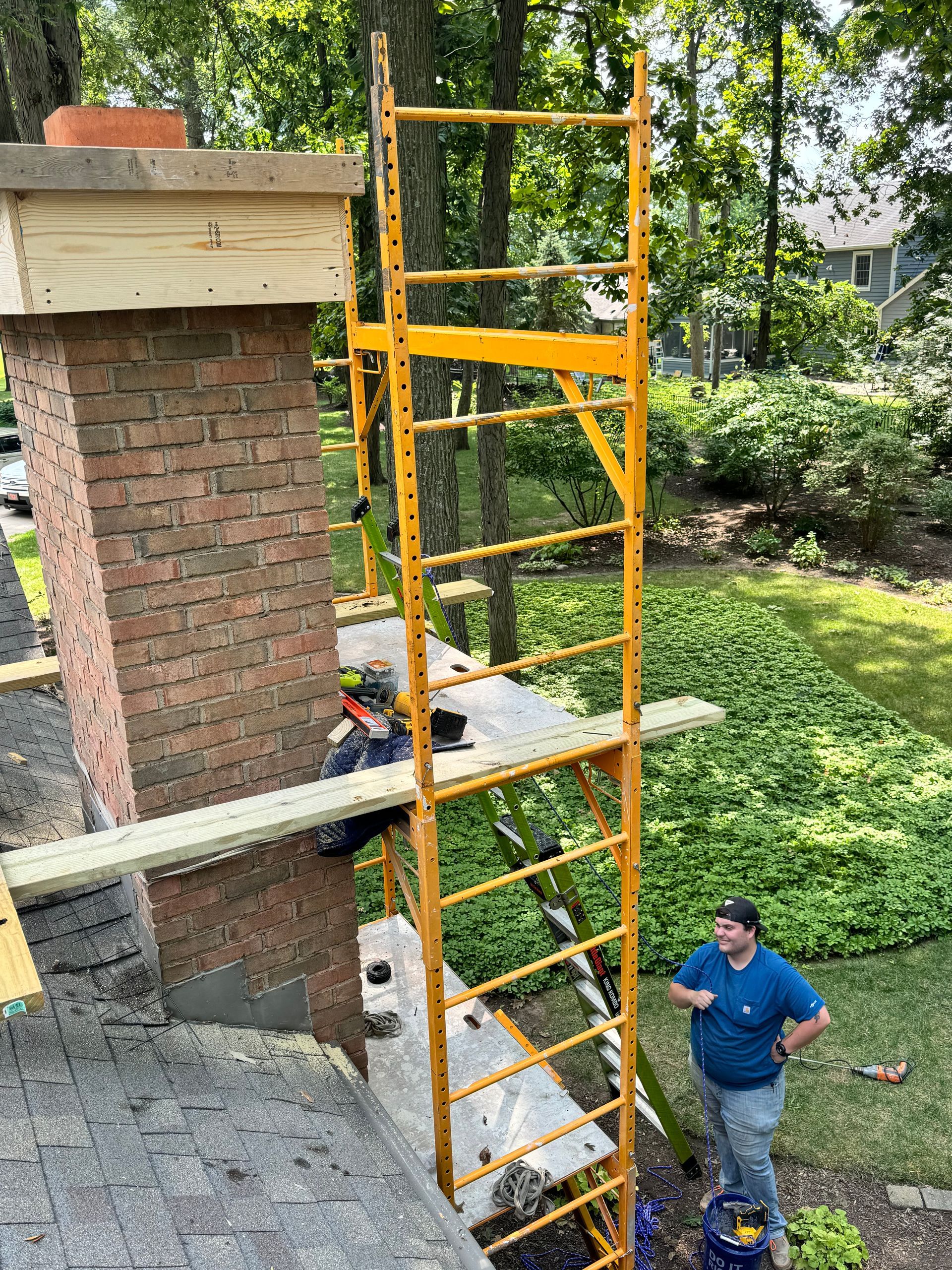 Orange ladder scaffold beside a brick chimney, with a person standing below on the lawn.