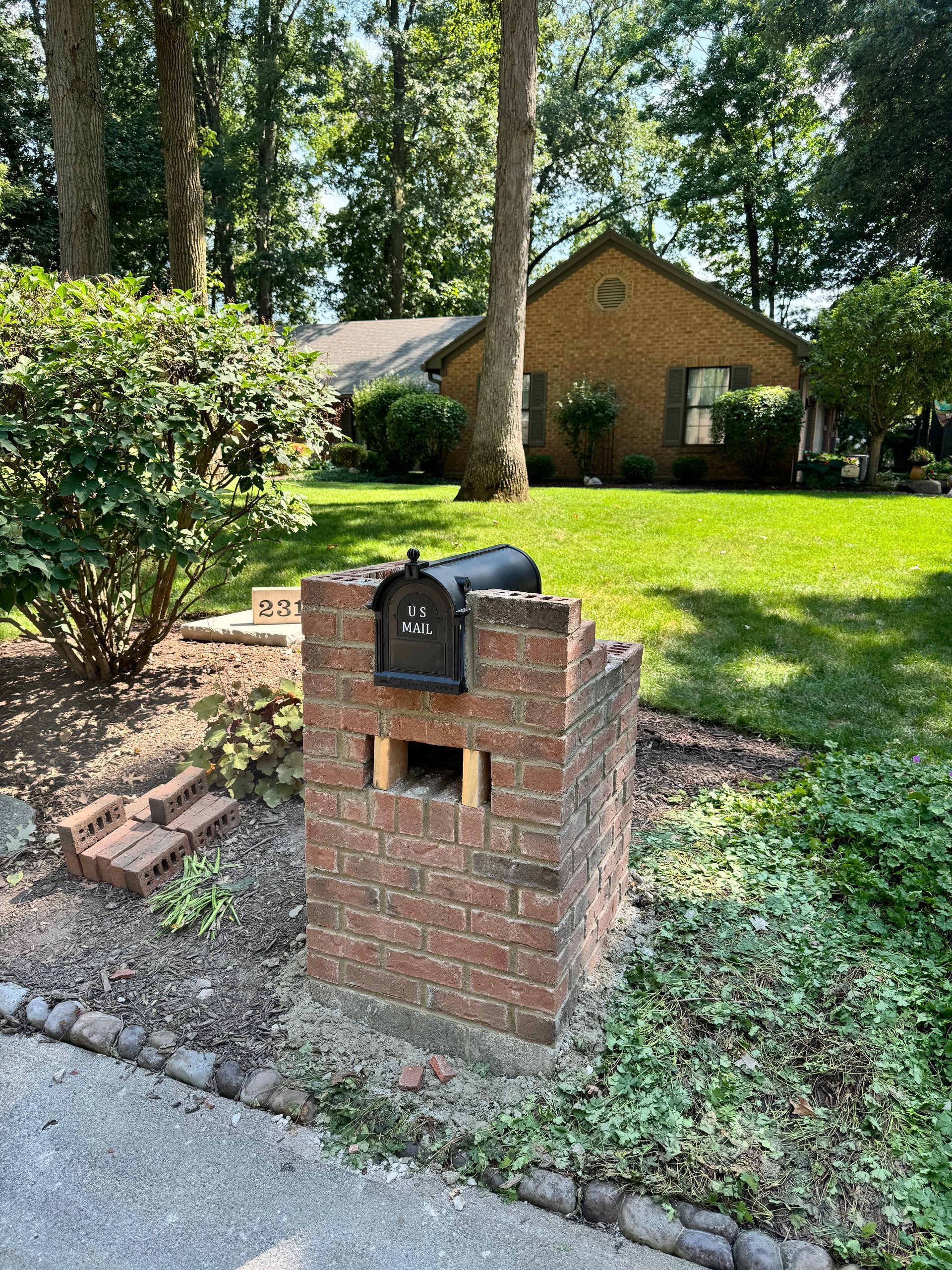 Brick mailbox in a front yard with trees, grass, and a house in the background