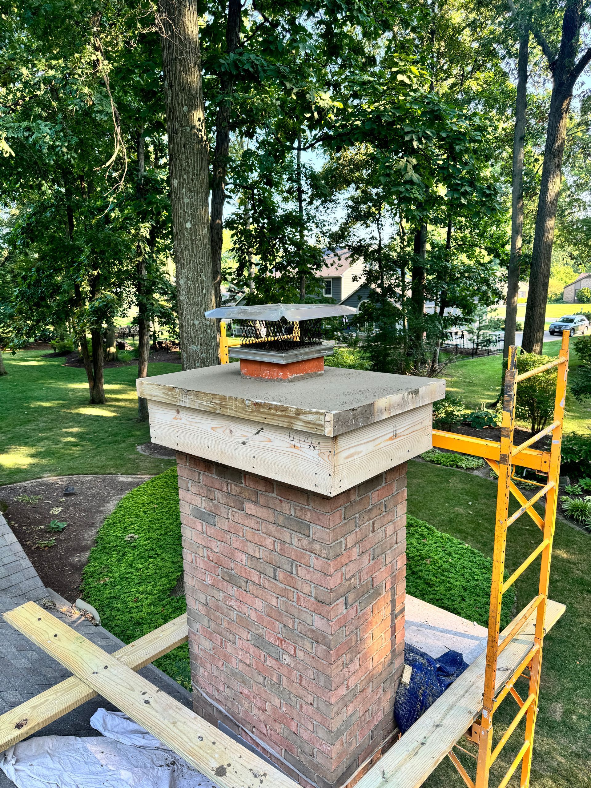 Brick chimney with a concrete cap in a wooded yard, surrounded by yellow scaffolding and construction materials