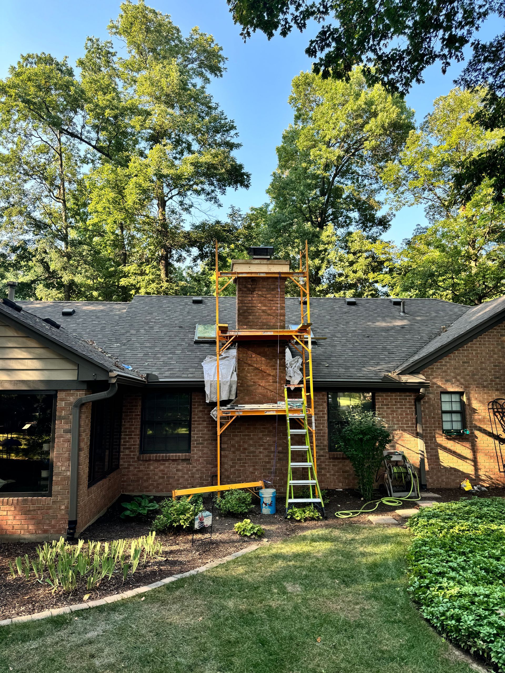 Brick house with a chimney under repair, scaffolding and ladder in front, trees and lawn around it