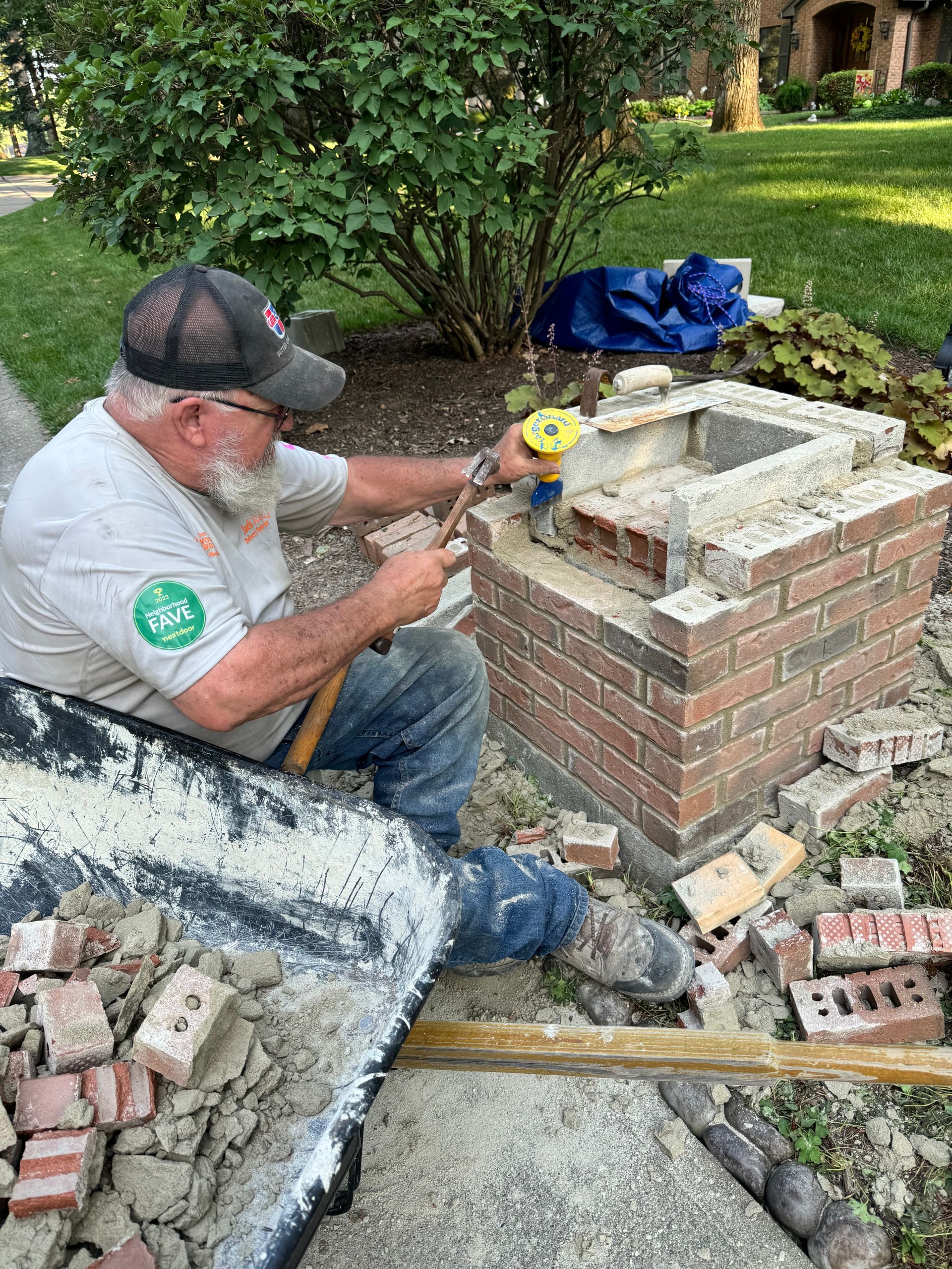 Worker laying bricks on a small outdoor brick structure in a garden, using tools and mortar.