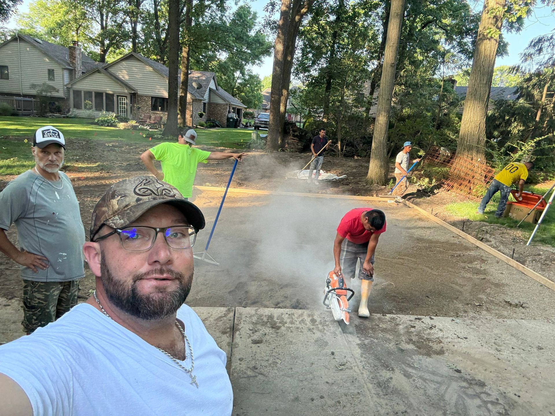 People paving a driveway; one man takes a selfie while others smooth wet concrete on a tree-lined street.